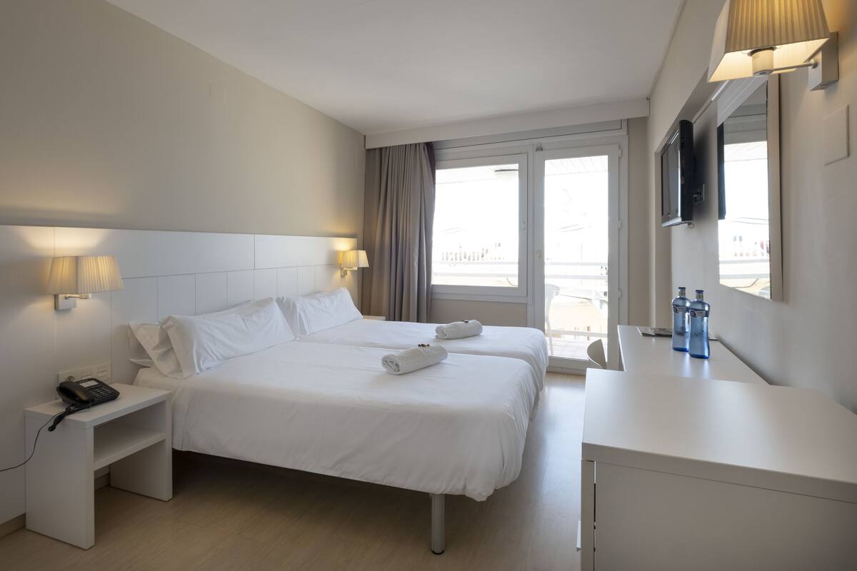 A hotel room with two beds, a bedside table with a phone, a desk with a TV, and a window with curtains.