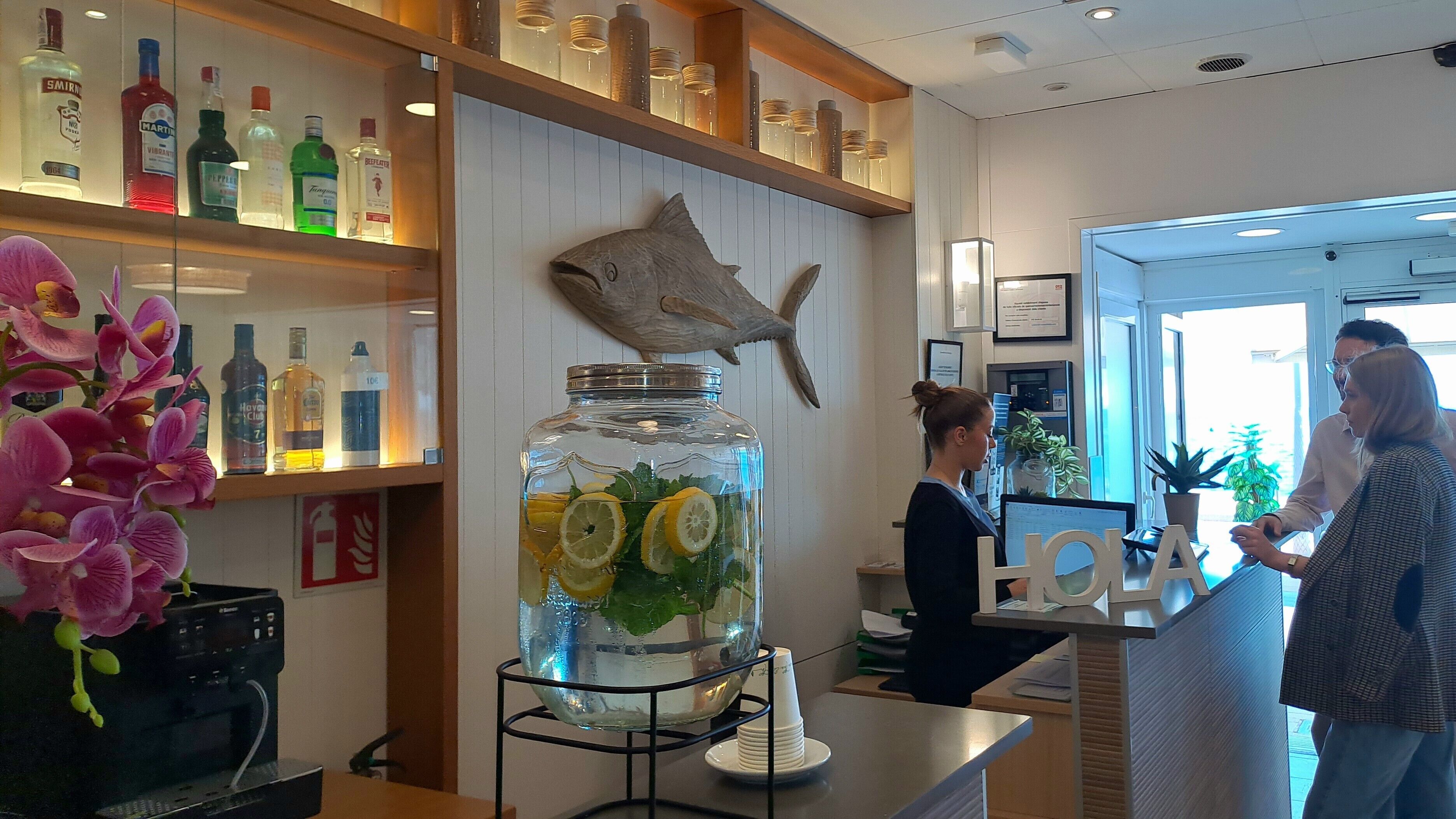 Un coin bar avec des étagères remplies de bouteilles d’alcool, une grande décoration en forme de poisson et un bocal contenant des citrons et de la menthe.