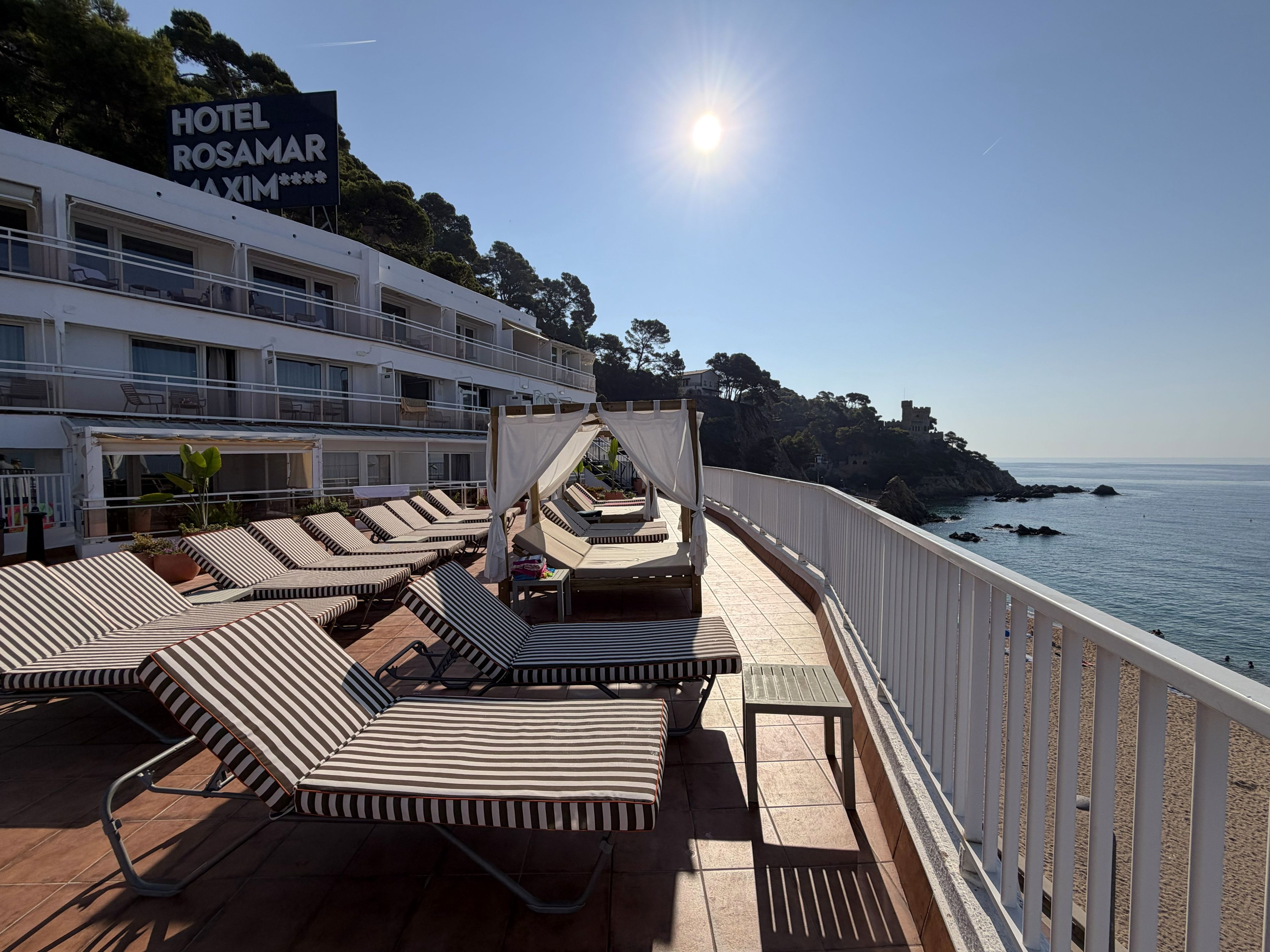 En hotellterrass med solstolar, en skylt för Hotel Rosamar Maxim och utsikt över havet.