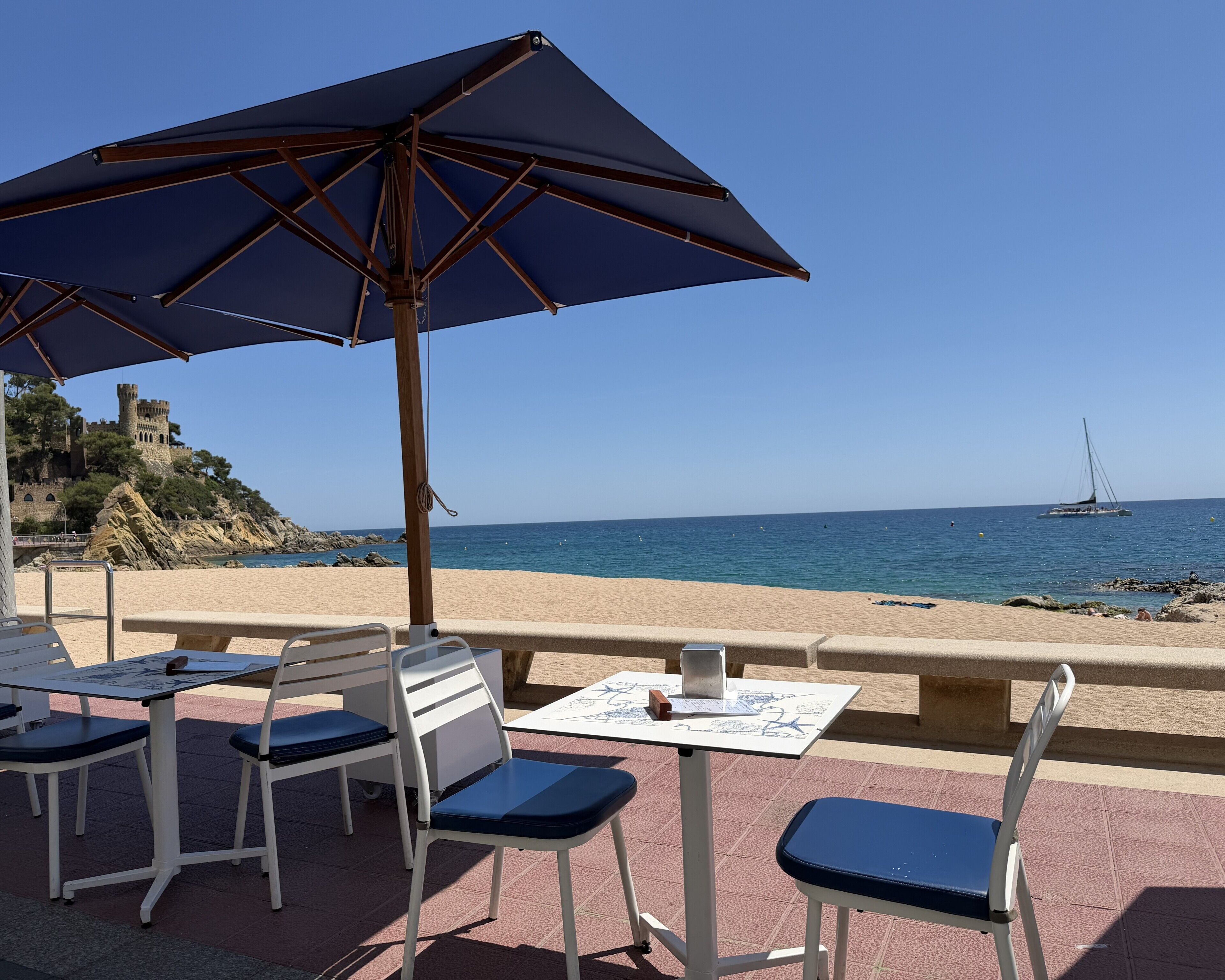 A beachside table with chairs and an umbrella, overlooking a sandy beach and a castle on a cliff.
