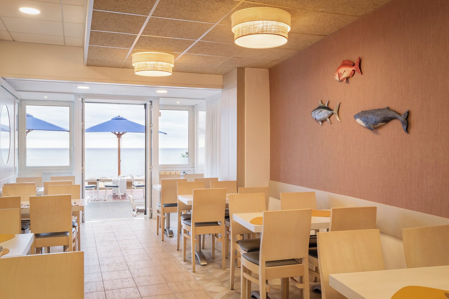 Ein Restaurant mit Holztischen und -stühlen, großen Fenstern und Meerblick.