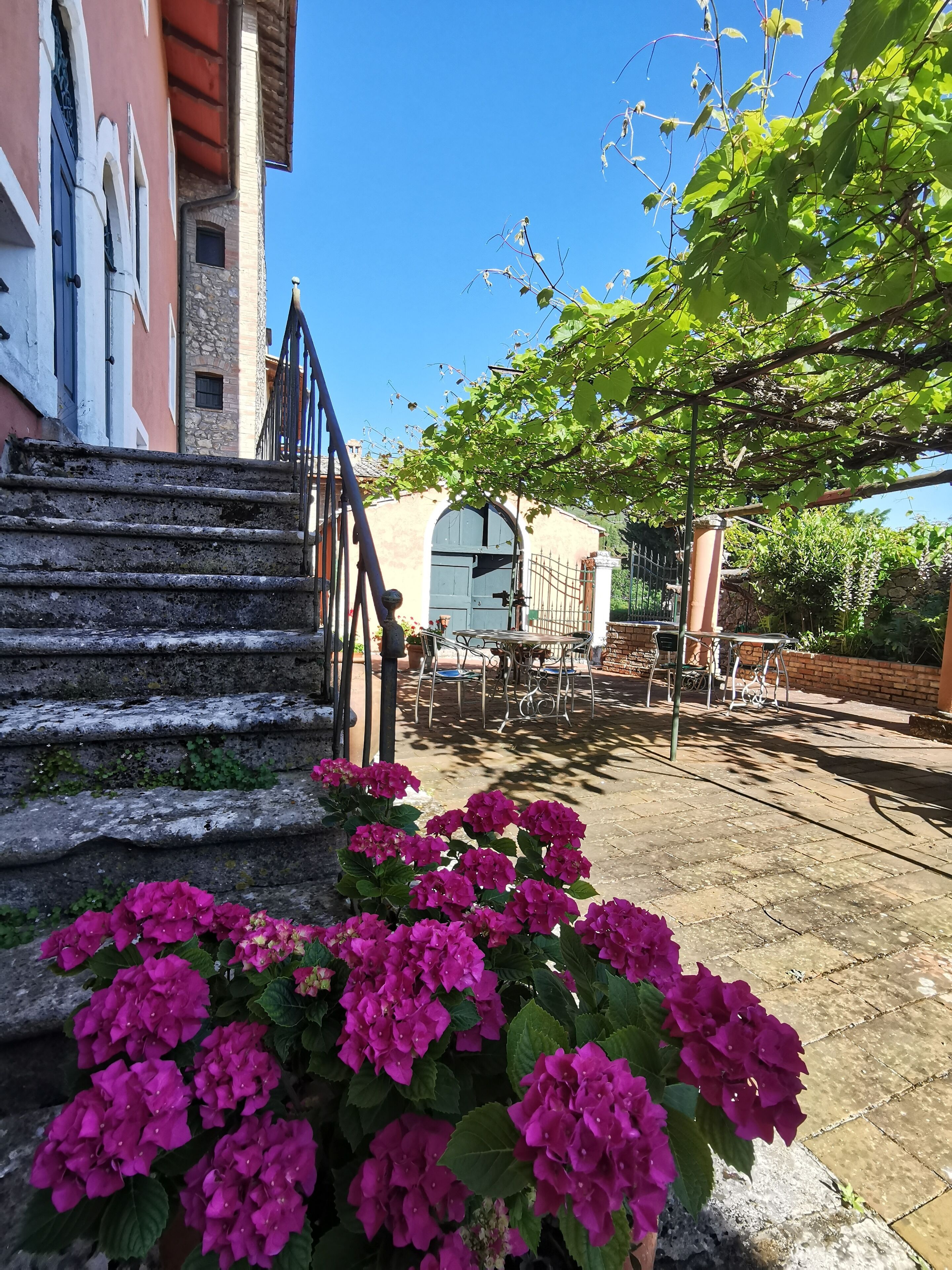 Un escalier en pierre mène à un bâtiment doté d’une porte blanche, avec une table et des chaises à l’extérieur, et des fleurs roses au premier plan.