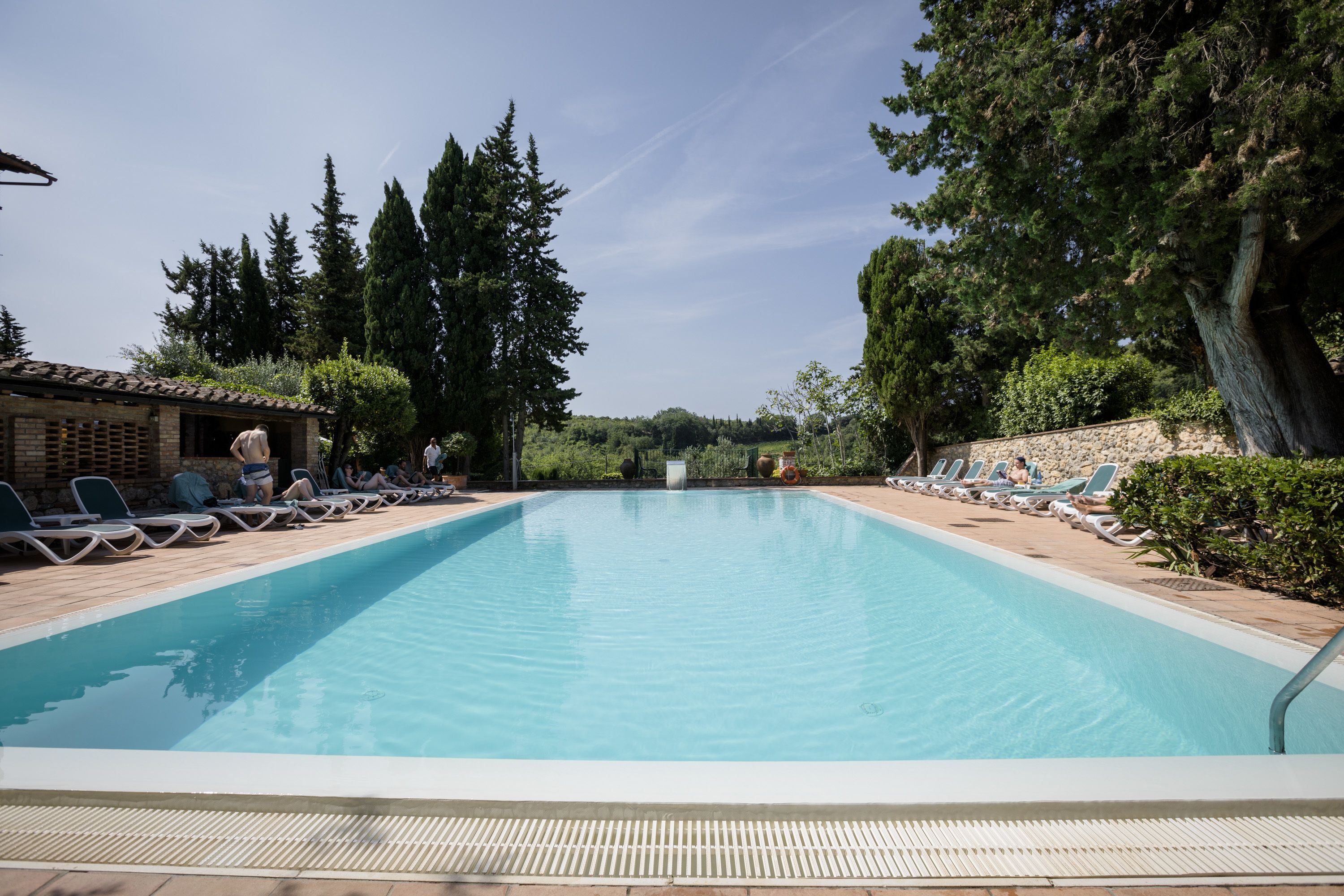 Piscina retangular com um deck revestido, rodeada por espreguiçadeiras e árvores.