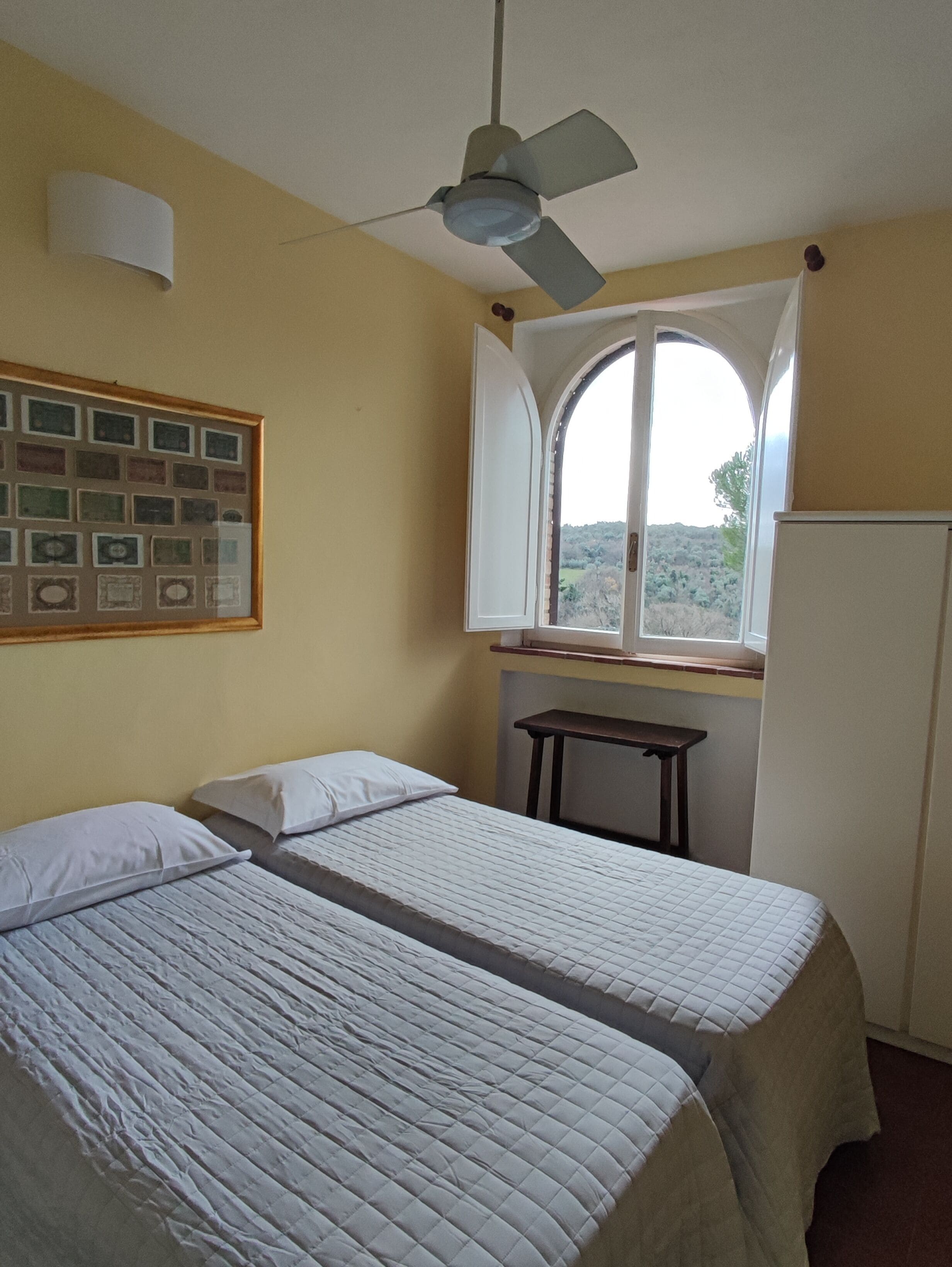 Habitació amb dos llits, un ventilador de sostre, una finestra amb vista i un quadre penjat a la paret.
