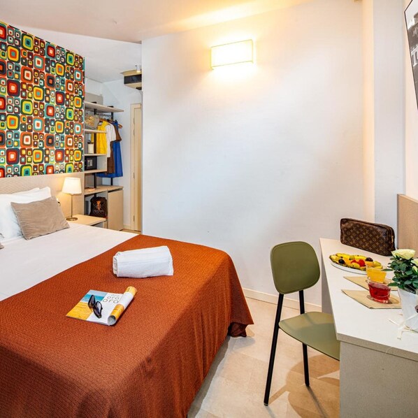 A hotel room with a bed, a desk with a chair, a small table with fruit and a drink, and a colorful wall.