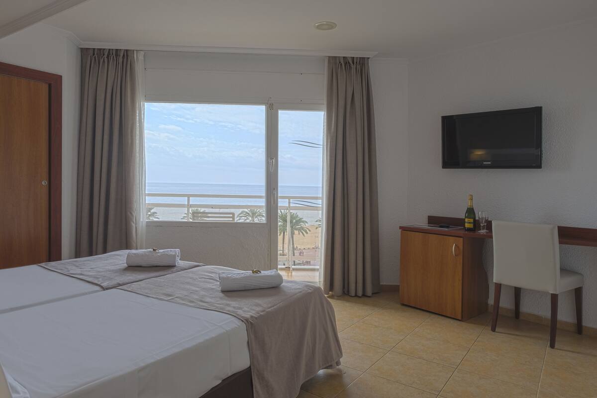 A hotel room with a bed, a desk with a bottle and glasses, a TV on the wall, and a view of the beach through the window.