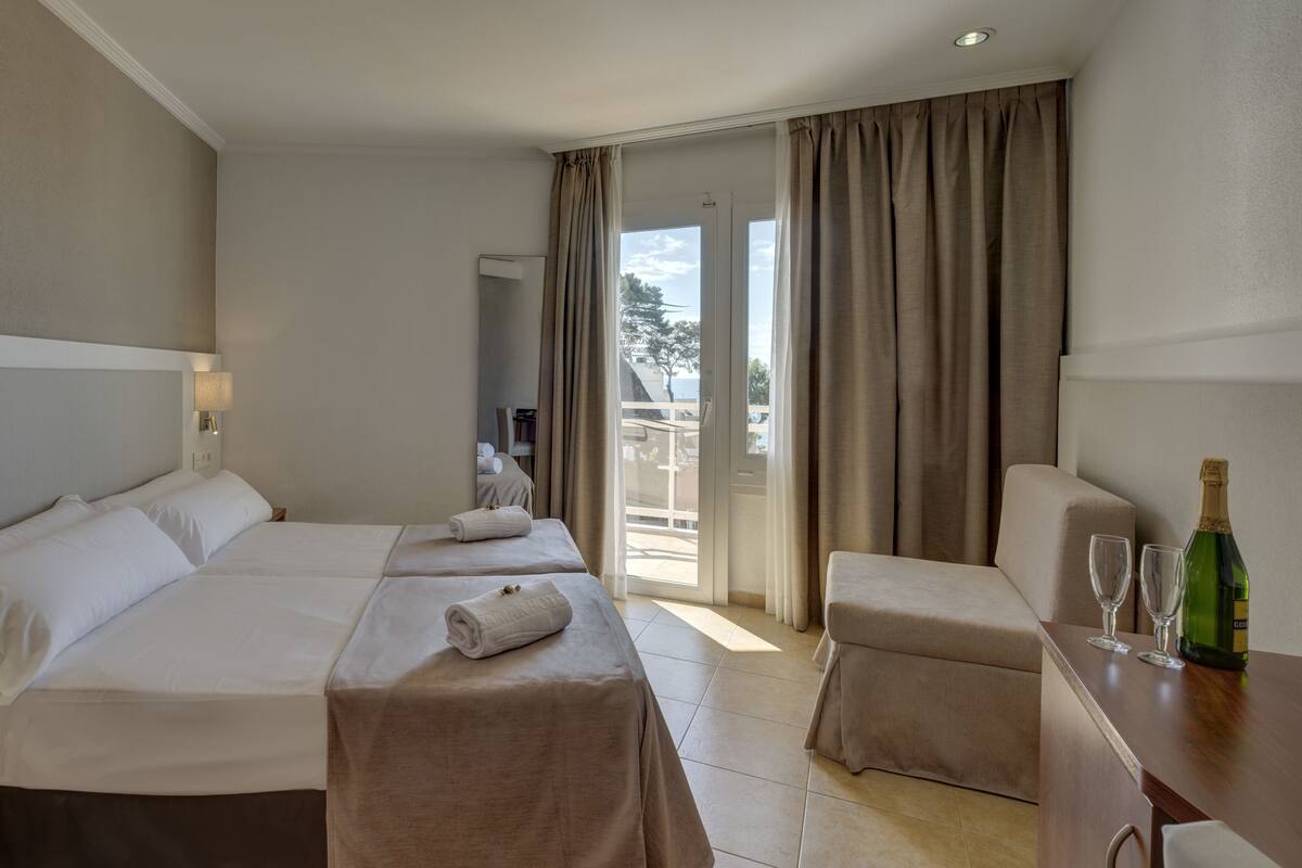 A hotel room with two beds, a desk, a chair, a minibar, and a balcony with curtains.