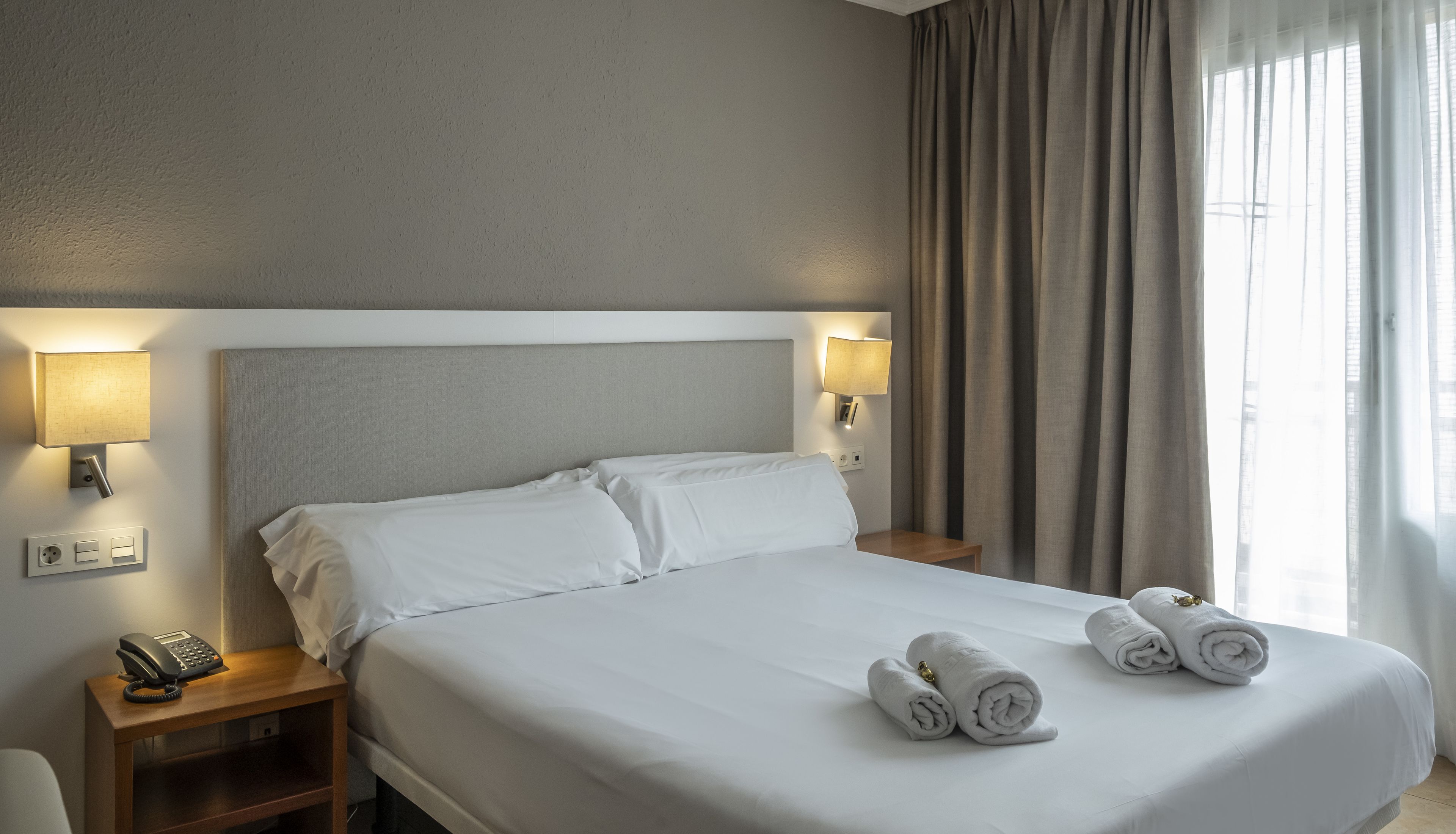 Ein Hotelzimmer mit einem großen Bett, zwei Nachttischlampen, einem Telefon und einem Fenster mit Vorhängen.