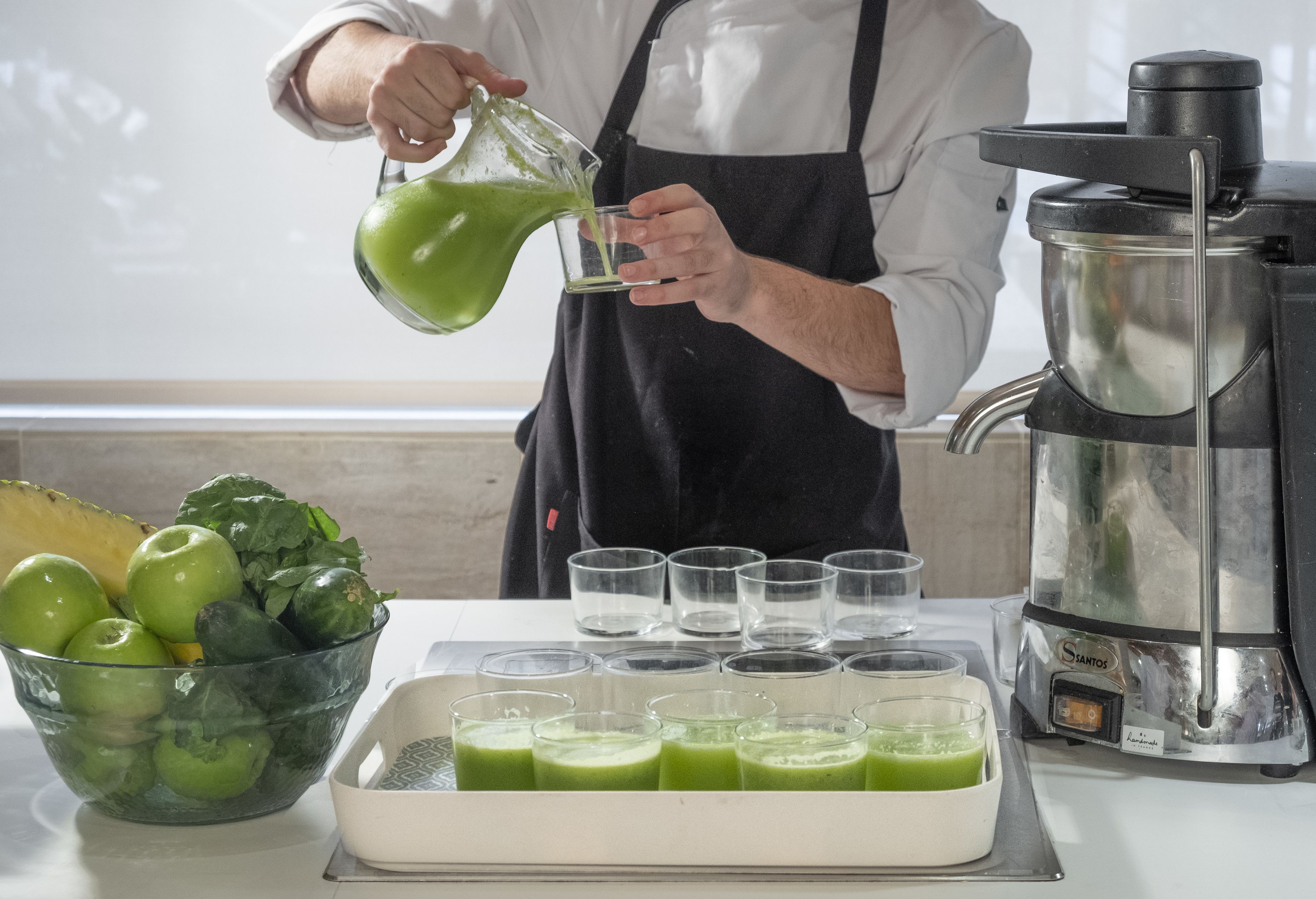 Una persona vertiendo un líquido verde en vasos, con una licuadora y un bol de manzanas verdes y verduras sobre una mesa.
