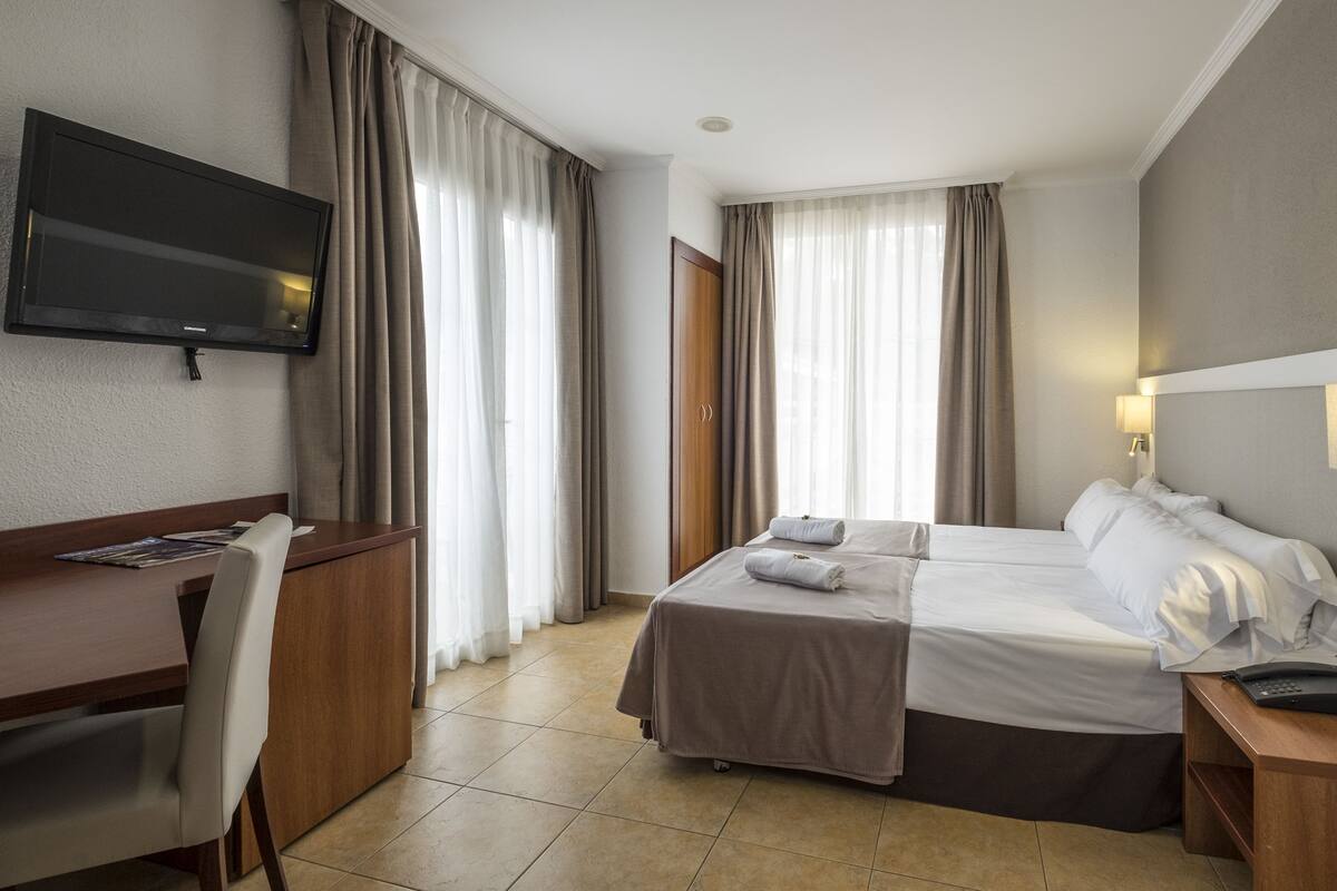 A hotel room with a bed, a desk, a chair, a TV, and a bedside table with a lamp.