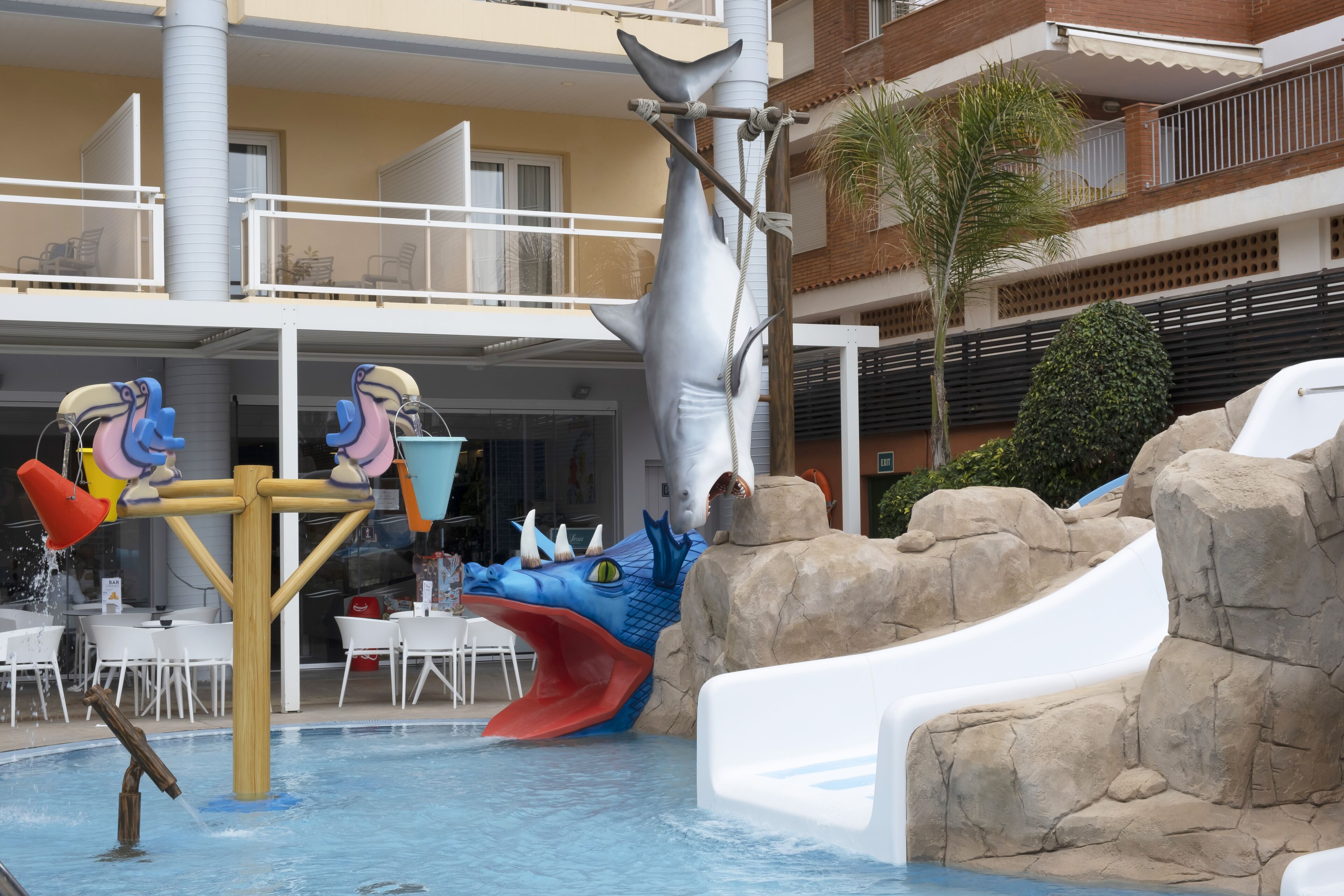 Un área de piscina con un tobogán acuático con temática de tiburón, una estructura de juego de delfines y un espacio de descanso con mesas y sillas.