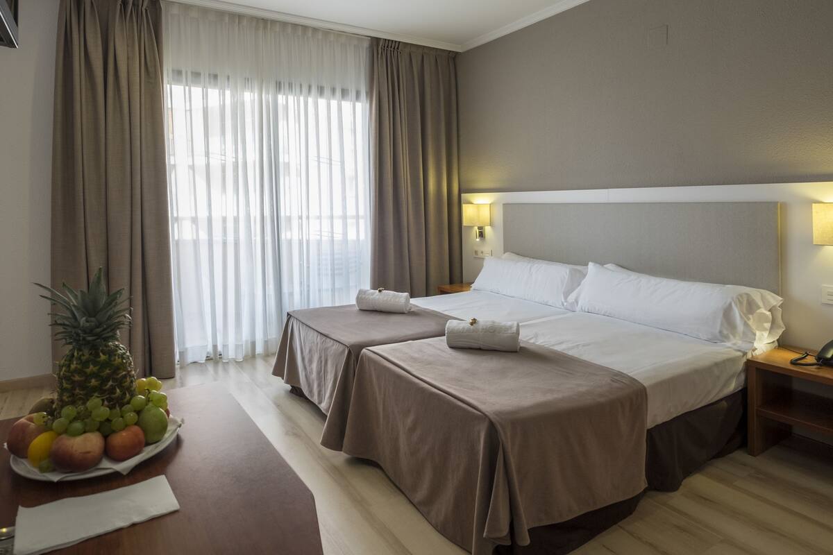 A hotel room with two beds, a fruit bowl on the table, and a view of the outdoors through sheer curtains.