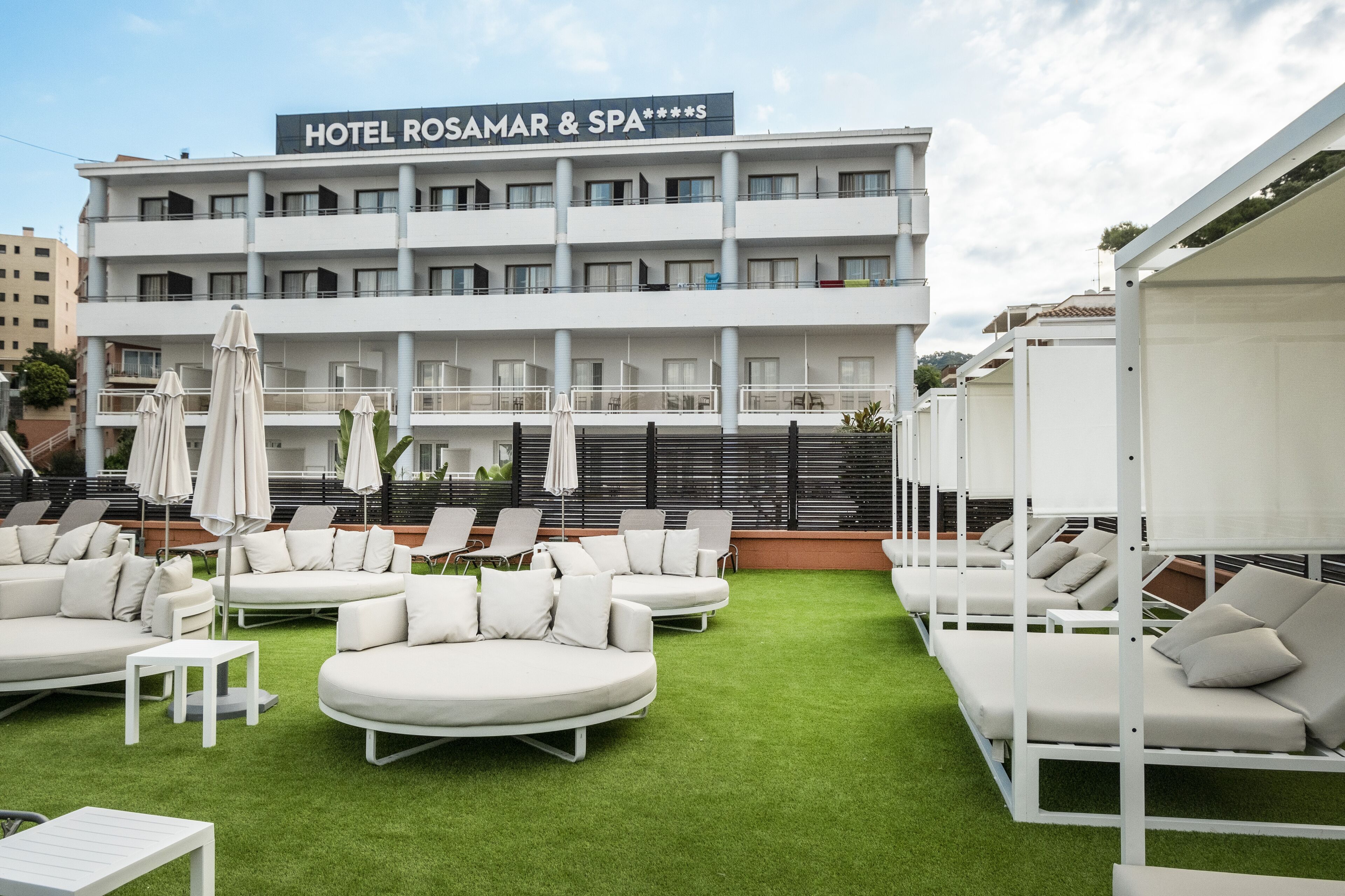A hotel with outdoor seating areas, including white sofas and umbrellas, and a sign reading "HOTEL ROSAMAR & SPA".