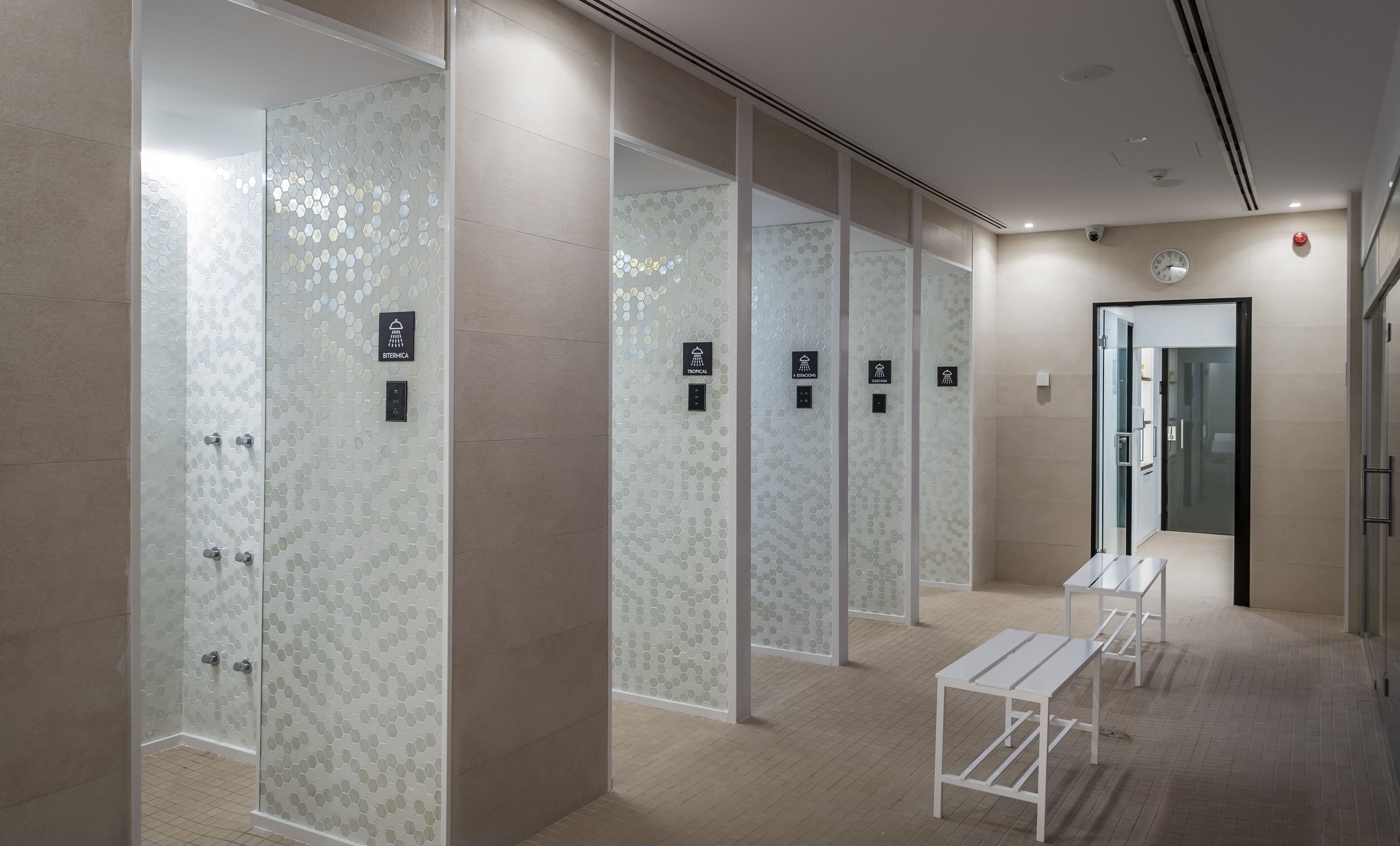 Une salle de bain moderne avec des douches à parois de verre, une horloge et un détecteur d’incendie au mur.