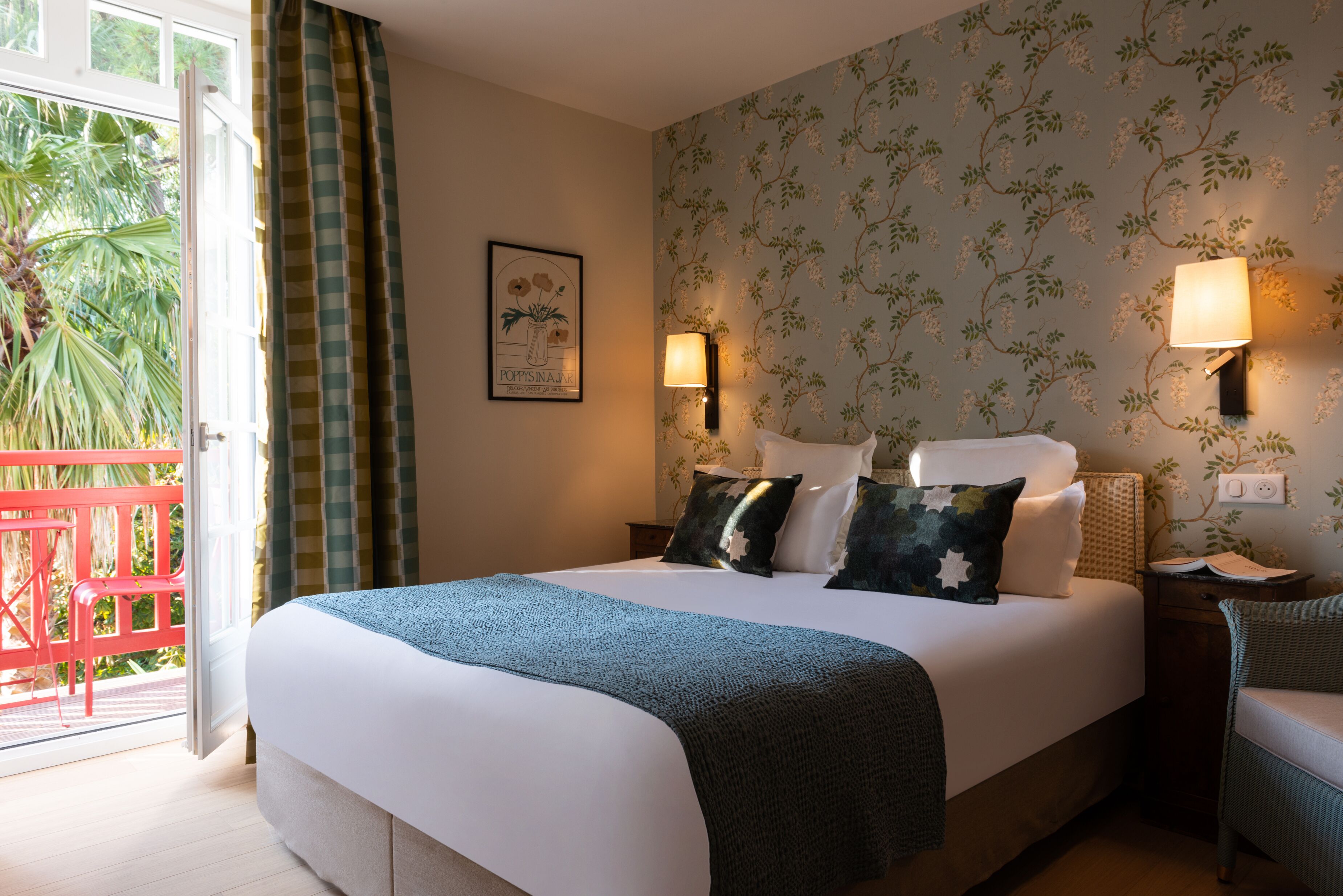 Un dormitorio con una cama, dos lámparas de noche, una mesita de noche, una silla, un ventanal con cortinas y una pared con papel tapiz floral.