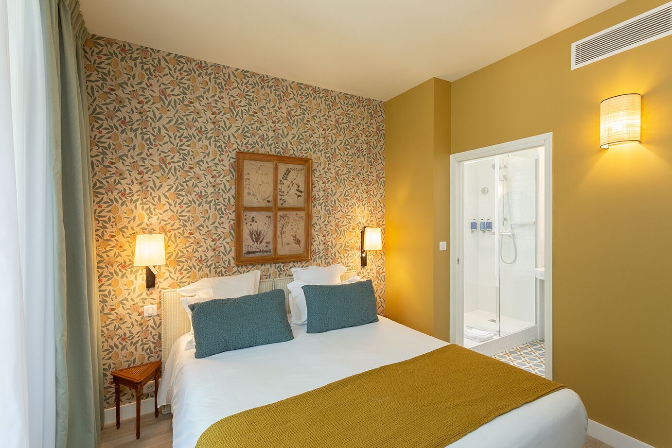 Habitación de hotel con cama, dos lámparas de pared, ducha y papel tapiz estampado con motivos florales.