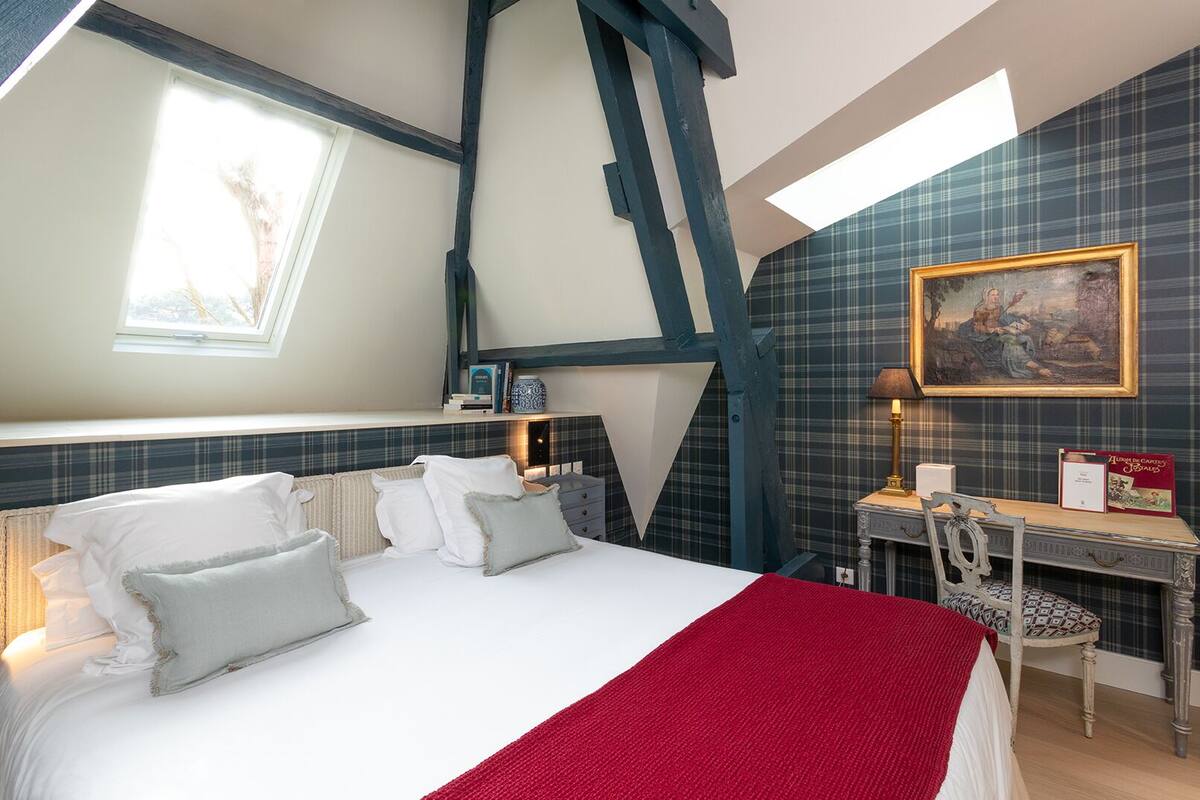 A bedroom with a bed, bedside tables, a desk, and a chair. There is a window and a painting on the wall.