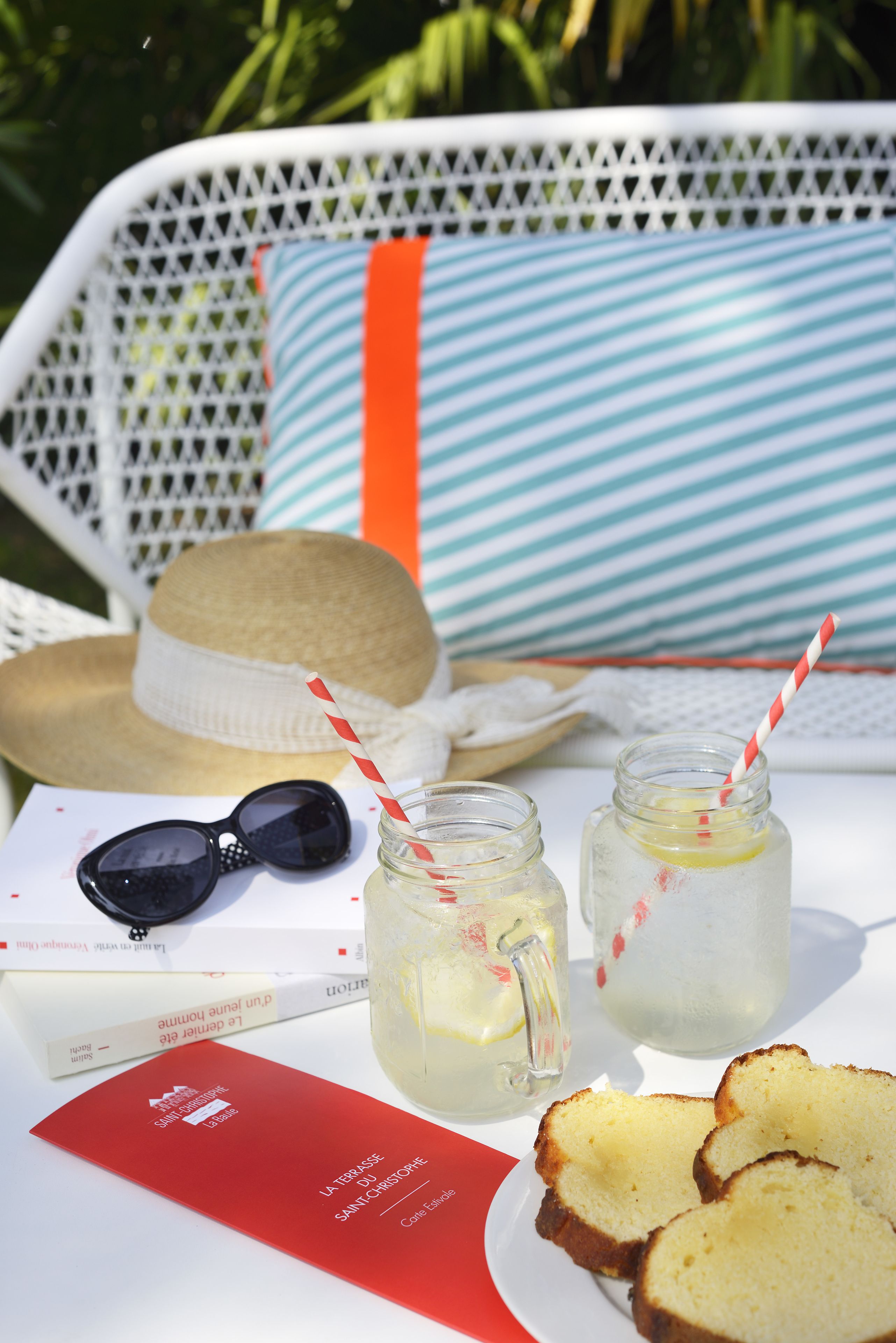 Una silla de mimbre con un cojín a rayas, un sombrero de paja, gafas de sol, un libro, dos frascos de vidrio con pajitas y rebanadas de pan sobre una mesa.