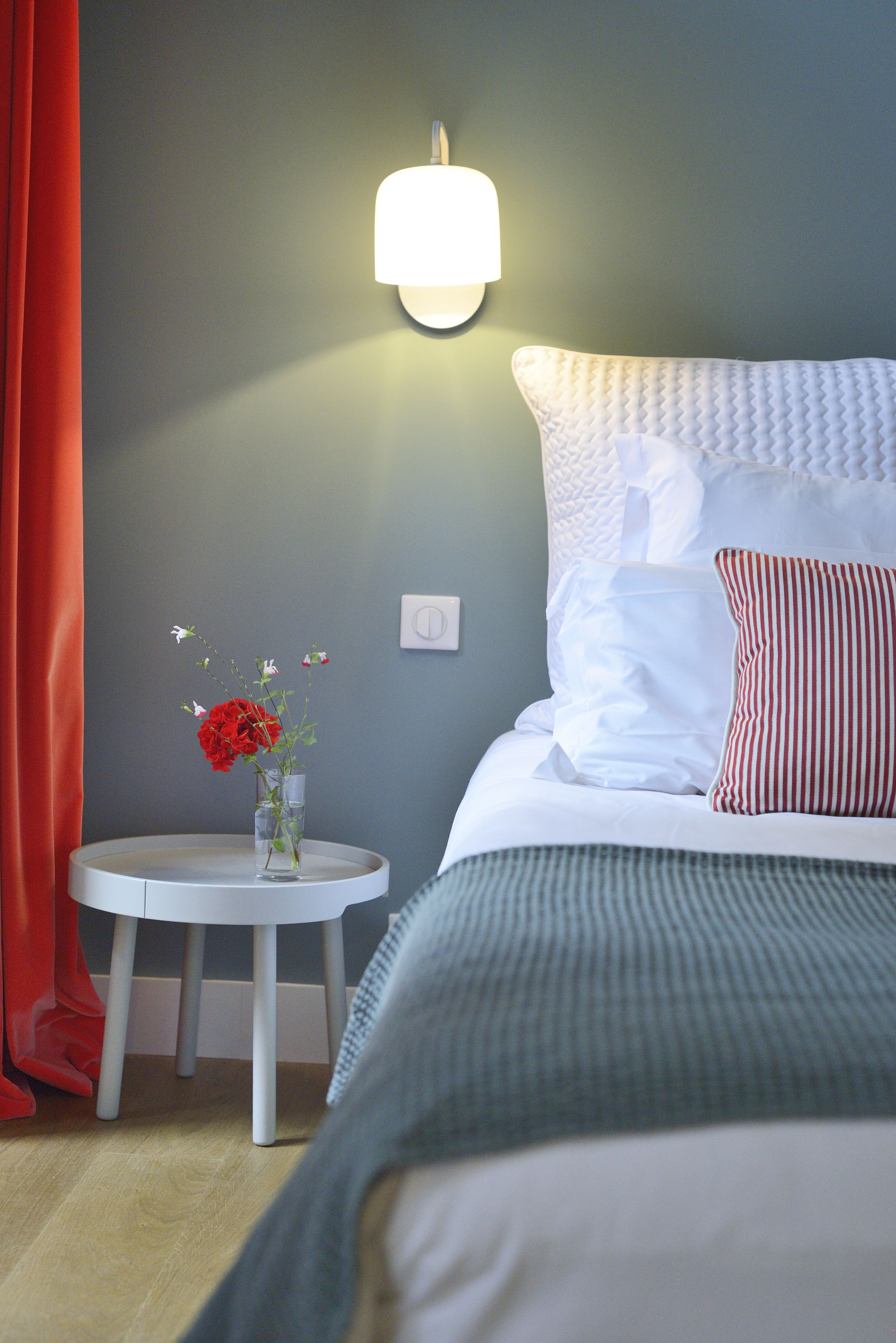 Un lit bien fait, avec des oreillers blancs et à rayures, une table de chevet ornée d’un vase de fleurs et un luminaire fixé au mur.
