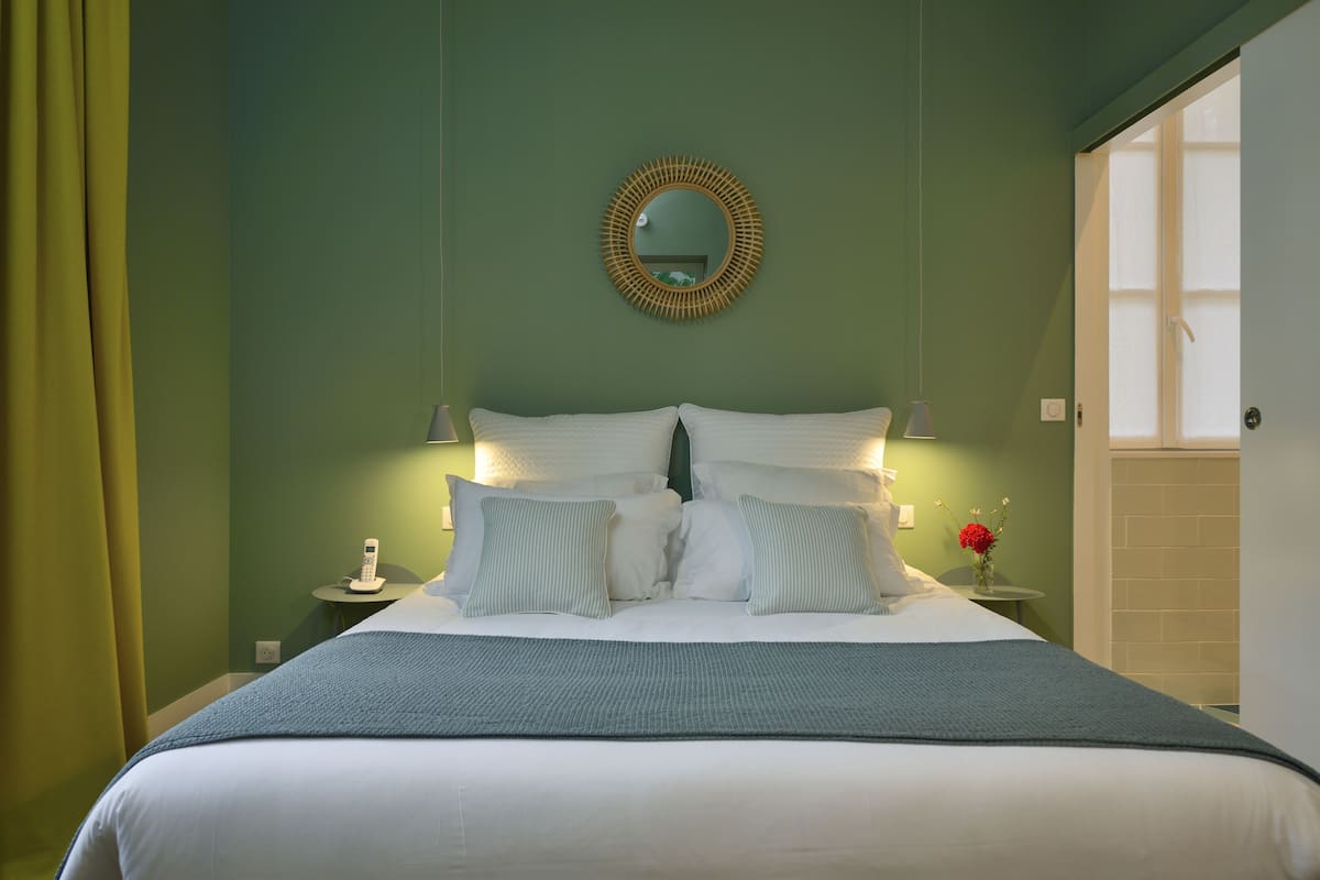 A neatly made bed with white linens and a blue throw blanket, a bedside table with a lamp and a vase of flowers, and a round mirror on the wall.