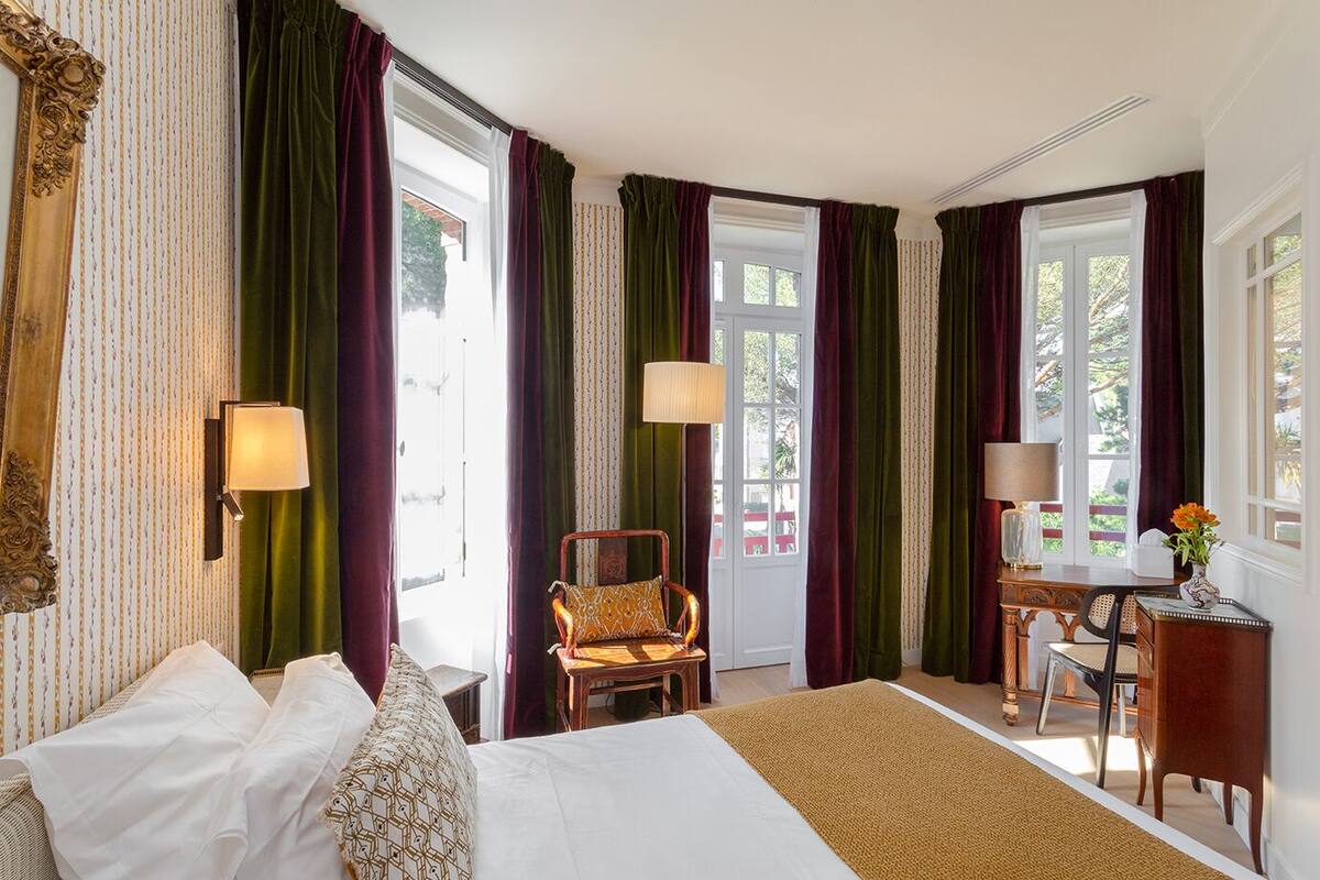 A hotel room with a bed, a desk, a chair, and large windows with curtains.