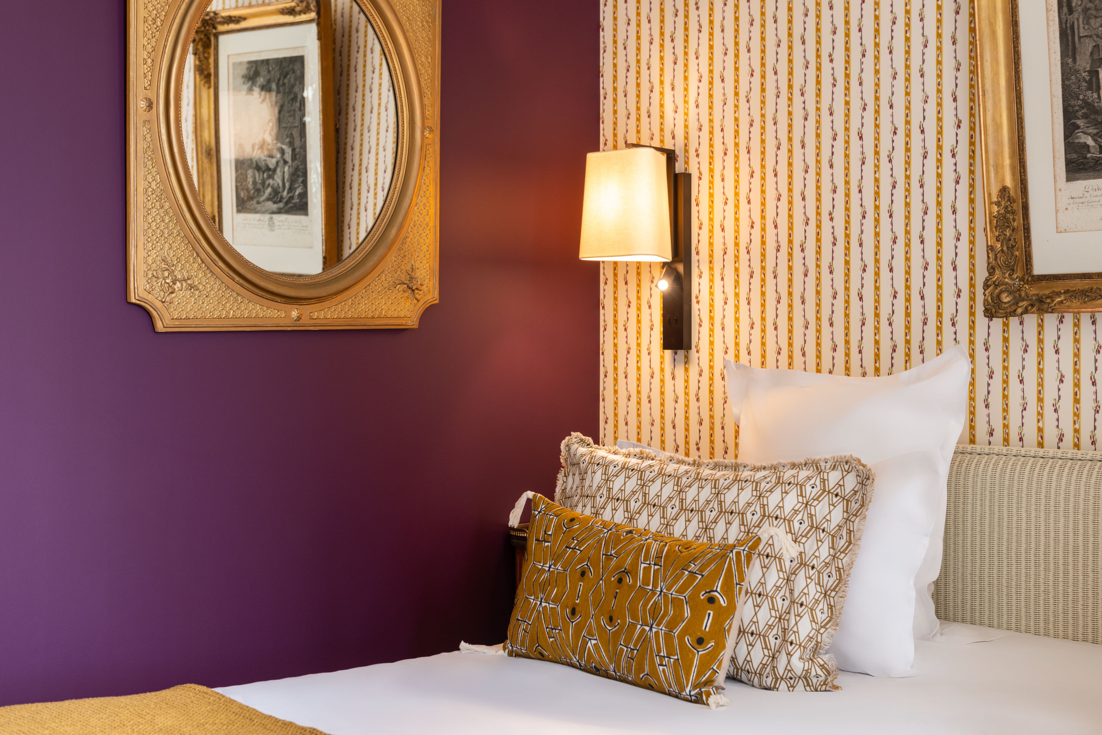 A hotel room with a purple wall, a framed picture, a wall-mounted lamp, and patterned bedding.