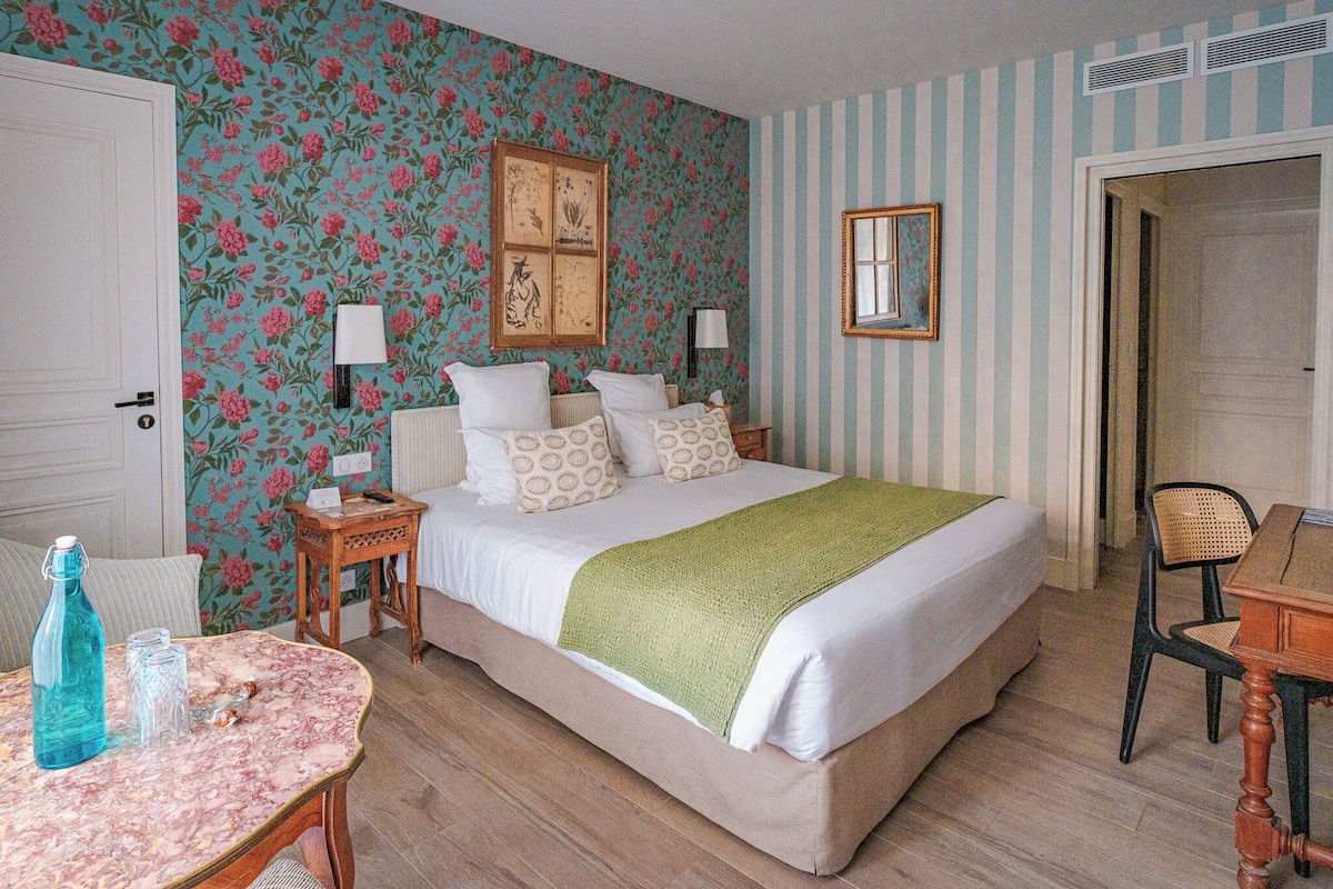 A bedroom with a floral wallpaper, a bed with patterned pillows, a green throw, a wooden nightstand, a table with a bottle and glasses, and a chair.