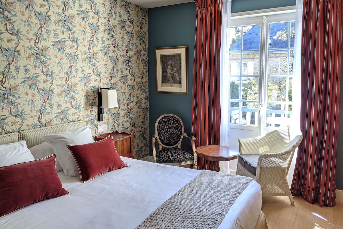 A hotel room with a large bed, a chair, a small table, and a window with curtains.