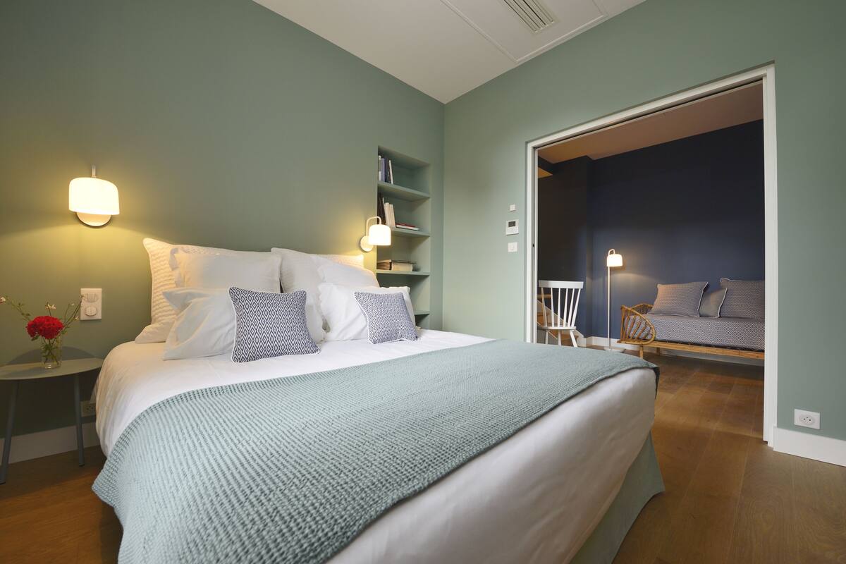A bedroom with a bed, bedside tables, a nightstand, a bookshelf, and a door leading to another room.