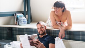 Un hombre leyendo un libro en una habitación con una ventana, una estantería y un jarrón decorativo.
