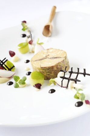 Um prato branco com carne, decorado com folhas verdes, frutas brancas e vermelhas, e grãos pretos.