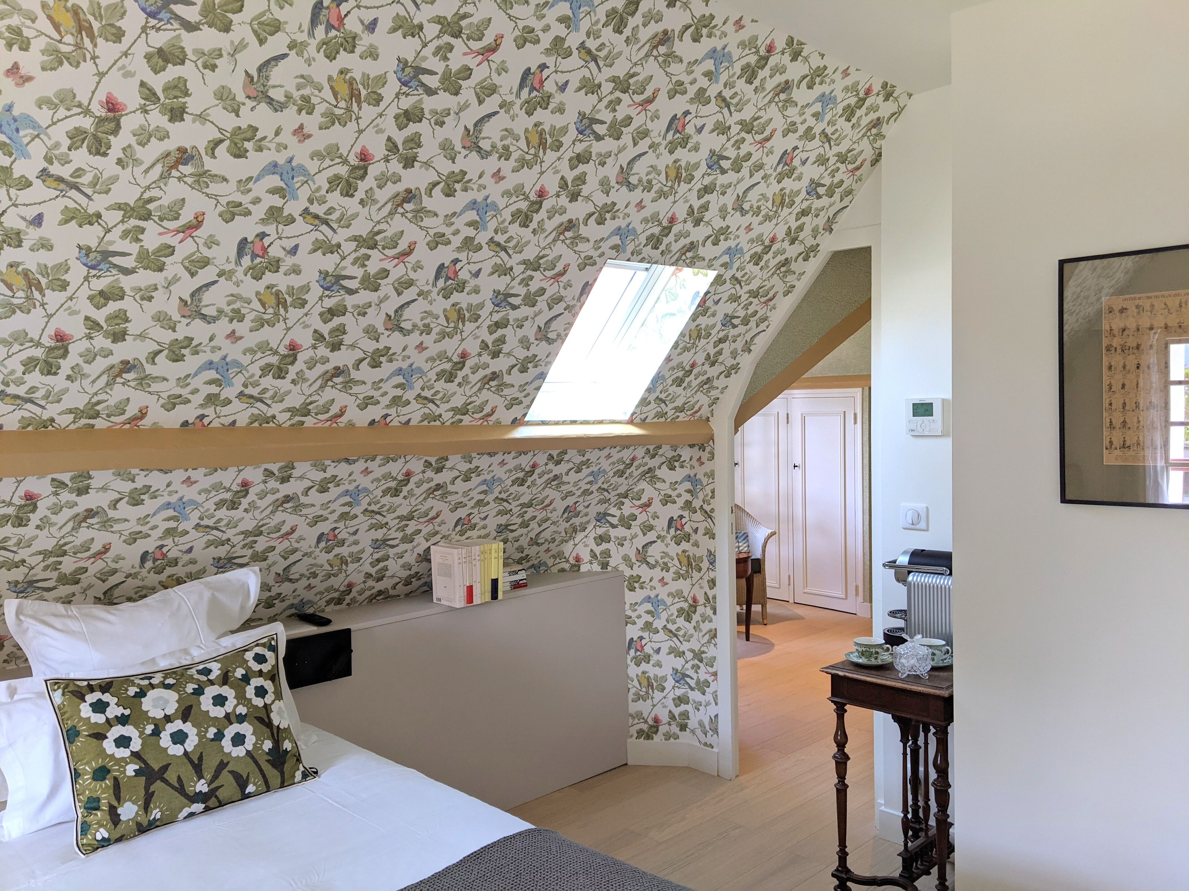 A bedroom with a sloped ceiling, a skylight, a bed with white bedding, a floral-patterned pillow, and a small table with a teapot.