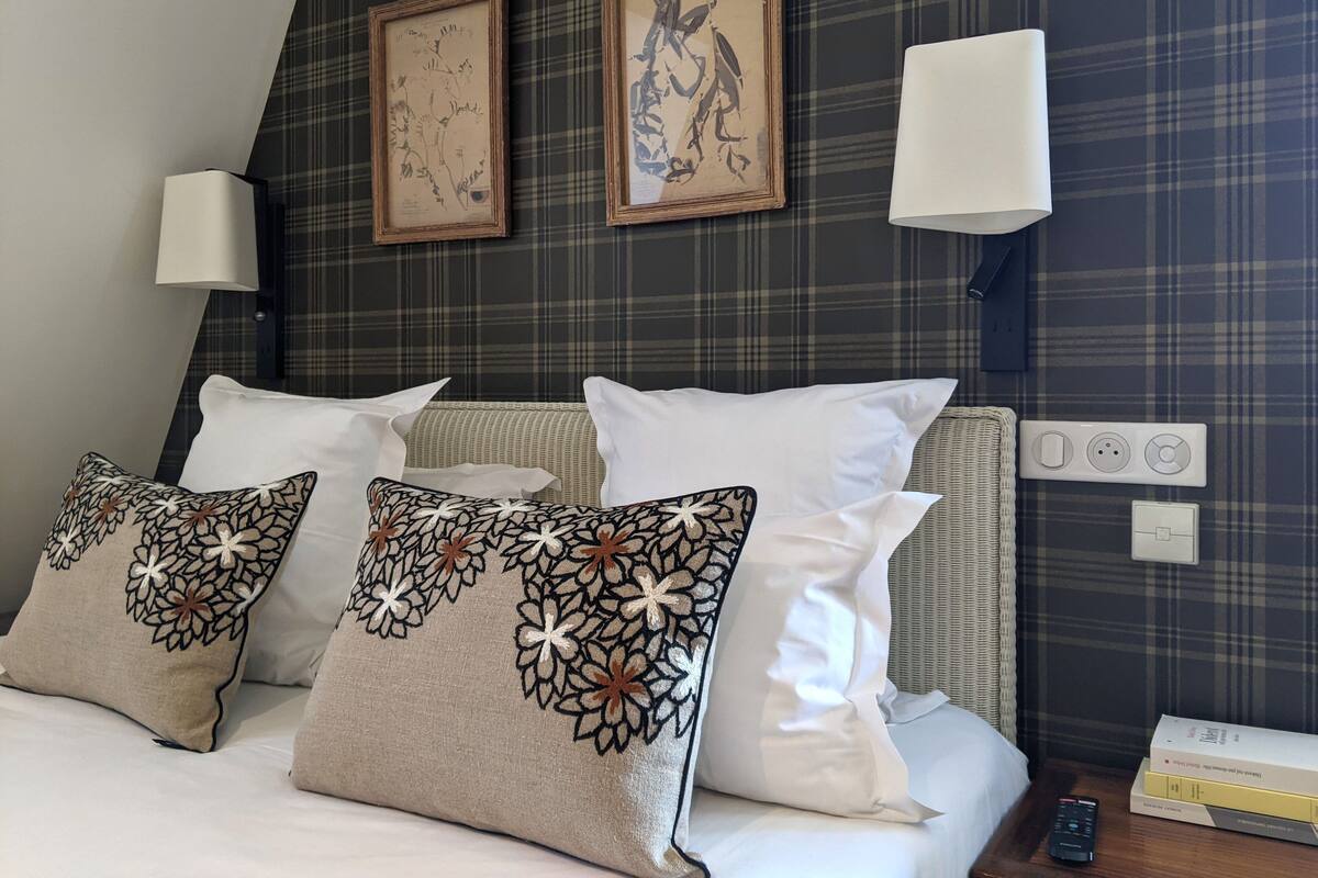 A hotel room with a bed, decorative pillows, two framed artworks, wall-mounted lamps, and a bedside table with a remote control and books.
