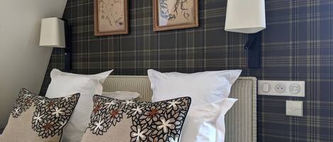 Een hotelkamer met een bed, decoratieve kussens, twee ingelijste kunstwerken, wandlampen en een nachtkastje met een afstandsbediening en boeken.