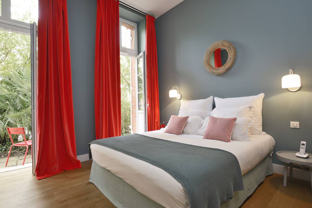 A bedroom with a bed, red curtains, a mirror, and a bedside table with a lamp.