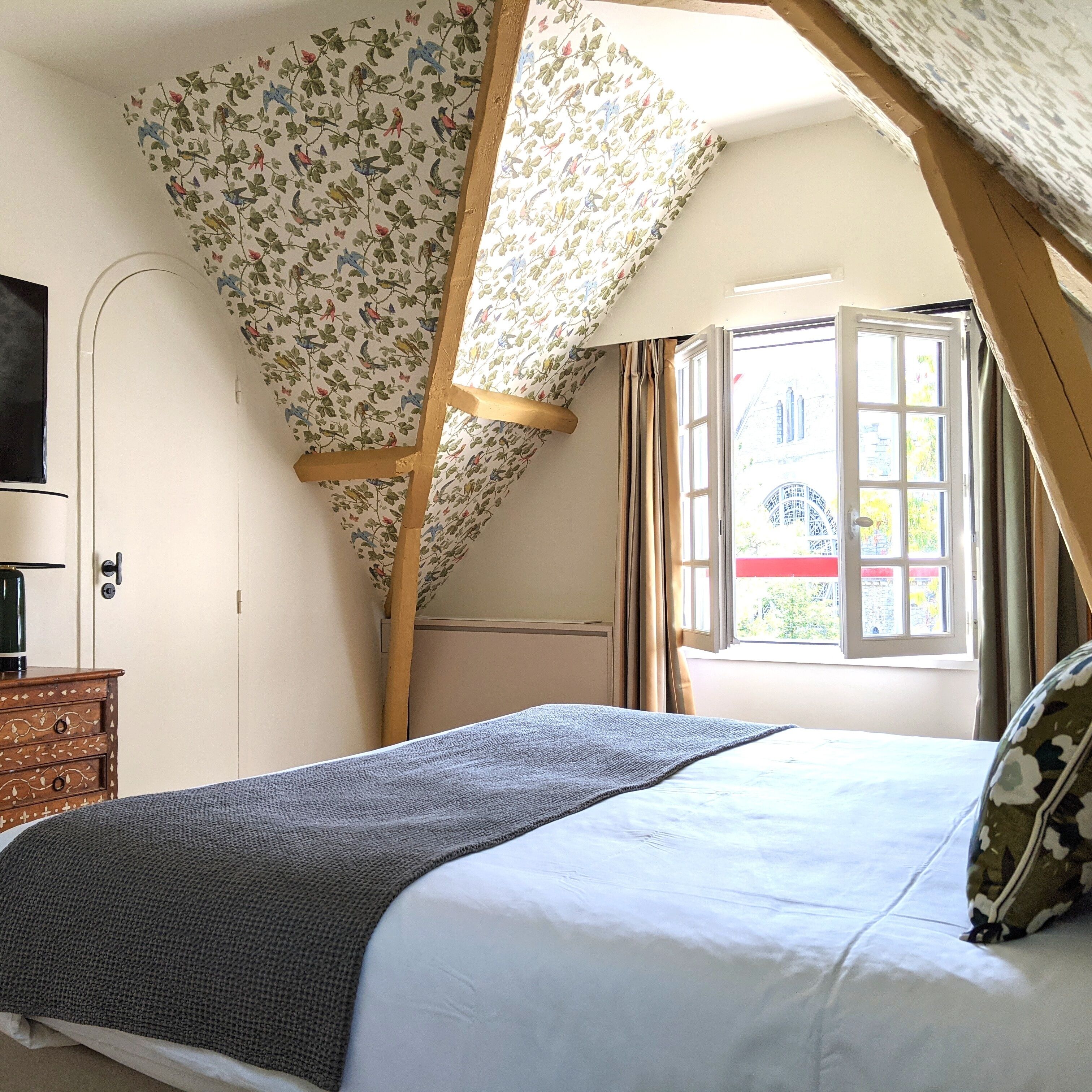 Un dormitorio con techo inclinado, una cama con manta gris, un aparador de madera y una ventana con cortinas.