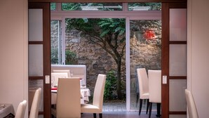 A dining area with white chairs and tables, a stone wall, and a tree visible through glass doors. - The Originals City, Hôtel Les Thermes de l'Avenue (Dax)