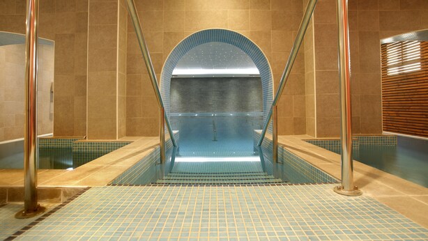 A modern indoor pool area with a clear glass wall, tiled floor, and metal railings. - The Originals City, Hôtel Les Thermes de l'Avenue (Dax)
