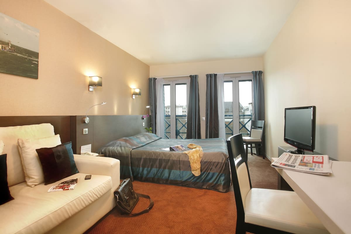 Ein Hotelzimmer mit Bett, einer Couch, einem Fernseher, einem Schreibtisch und einem Balkon mit Vorhängen.