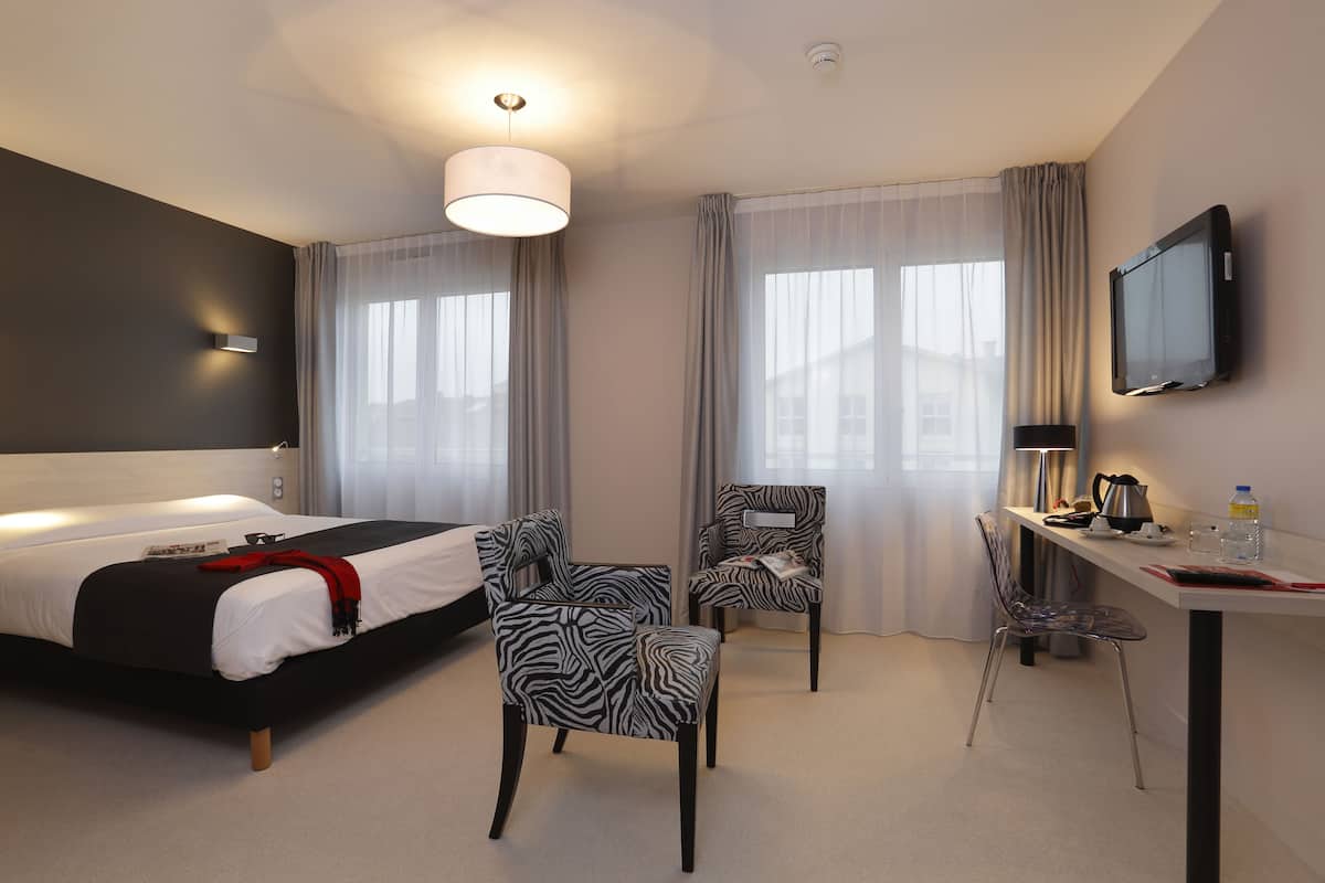 Ein modernes Hotelzimmer mit einem Bett, zwei Sessel, einem Schreibtisch mit Lampe und einem Fernseher.