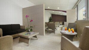 A modern lounge area with wicker furniture, a black sofa, and a coffee table with magazines. There are two wicker chairs and a small table with drinks. - The Originals City, Hôtel Les Thermes de l'Avenue (Dax)