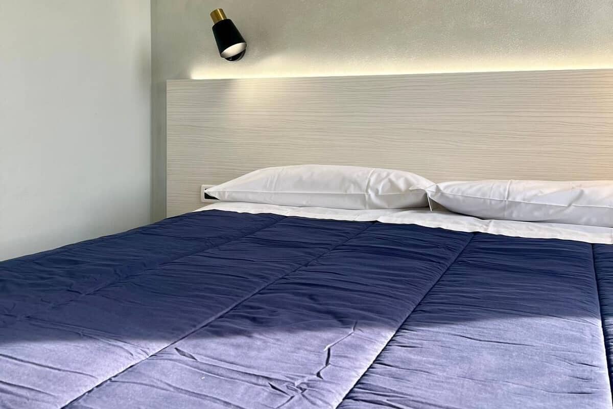 Una cama bien hecha con una colcha azul oscuro y almohadas blancas, un cabecero de color claro y una lámpara de noche pequeña.