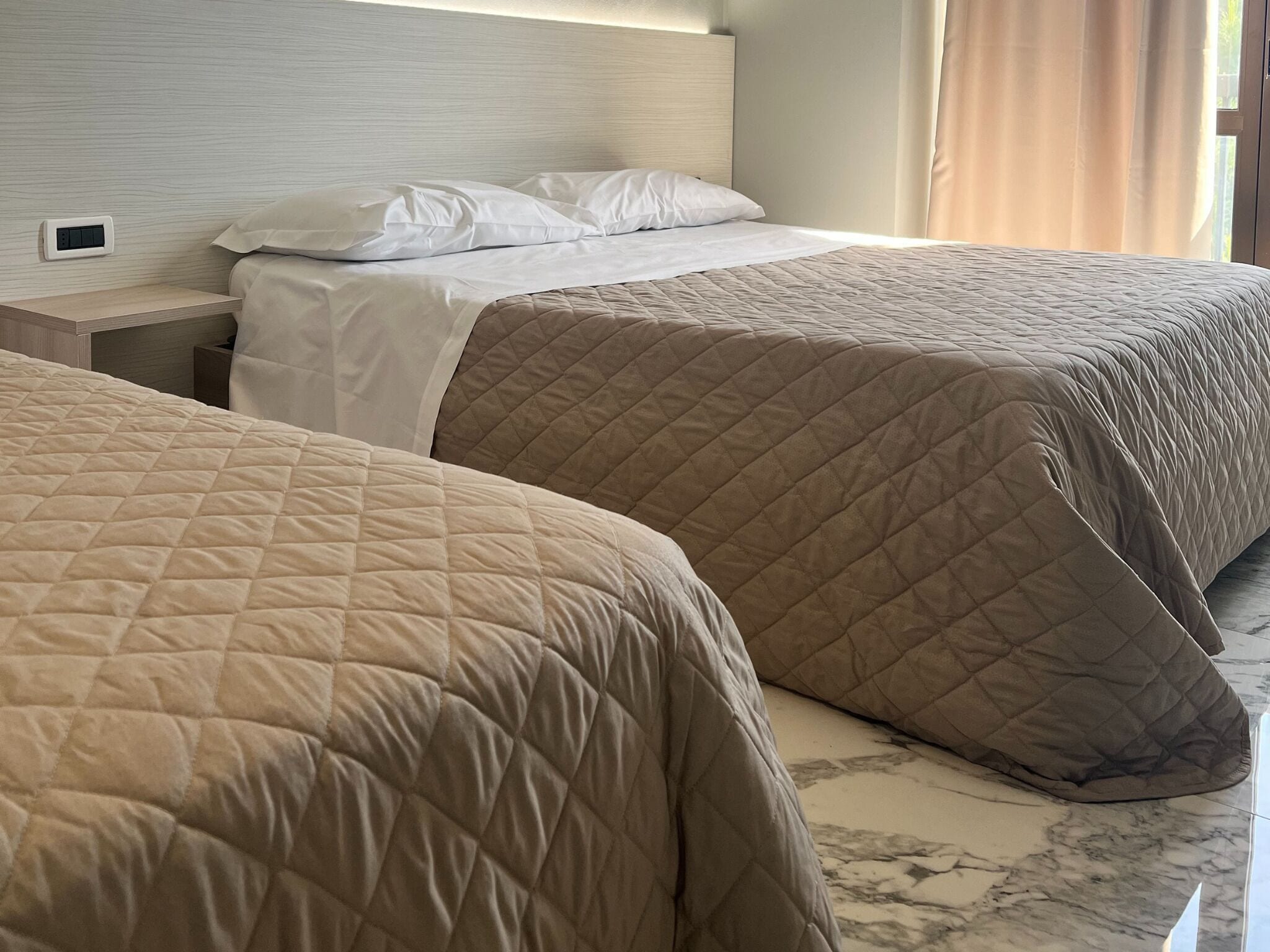 Habitación de hotel con dos camas, cada una con colcha acolchada y almohadas blancas. Se ve una mesita de noche con enchufe y lámpara. El piso es de mármol y hay una ventana con cortinas.