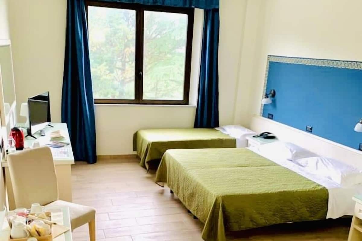 Habitación de hotel con dos camas individuales, un escritorio con computadora, una silla y un ventanal con cortinas.