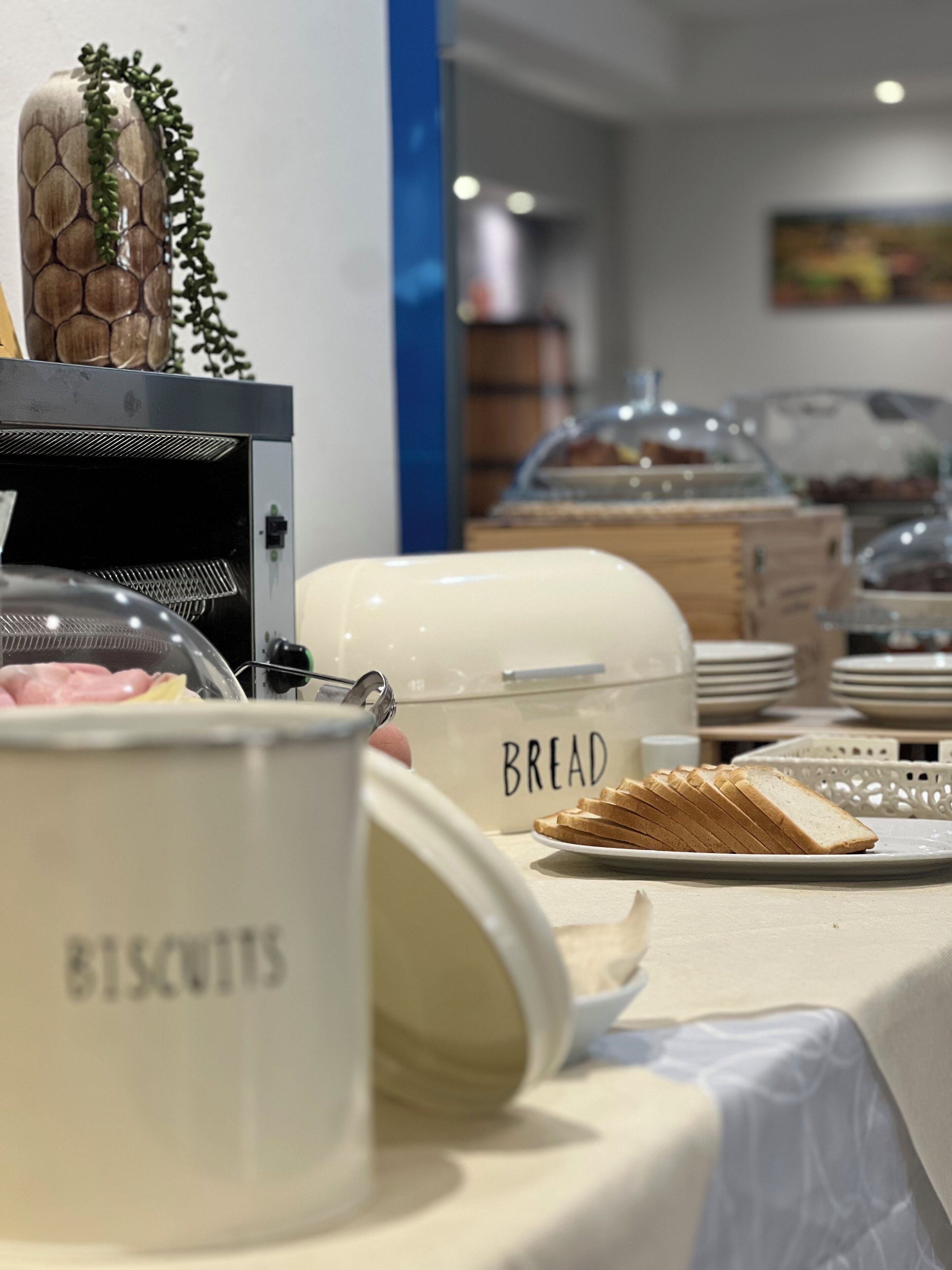 Montaje de bufé con recipientes etiquetados para pan y galletas, una campana de cristal sobre una bandeja de comida y un plato de pan en rodajas.