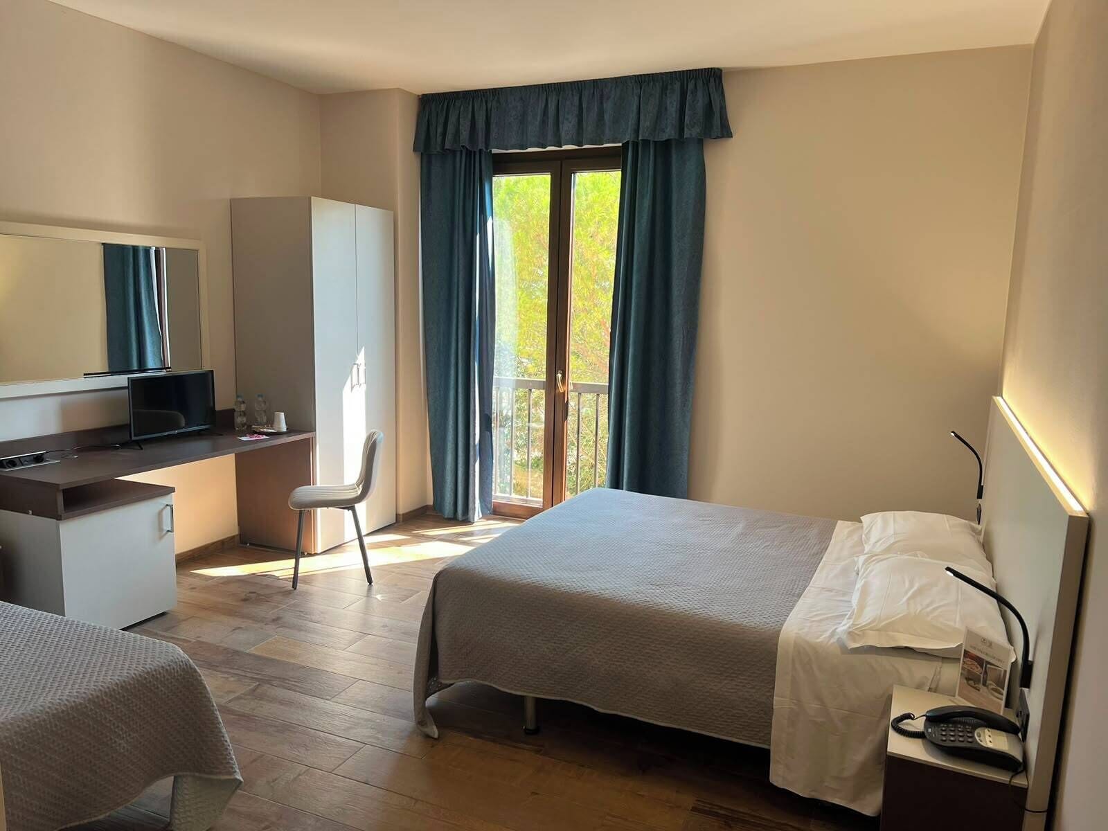 Habitación de hotel con cama, escritorio, silla, armario y balcón con cortinas.