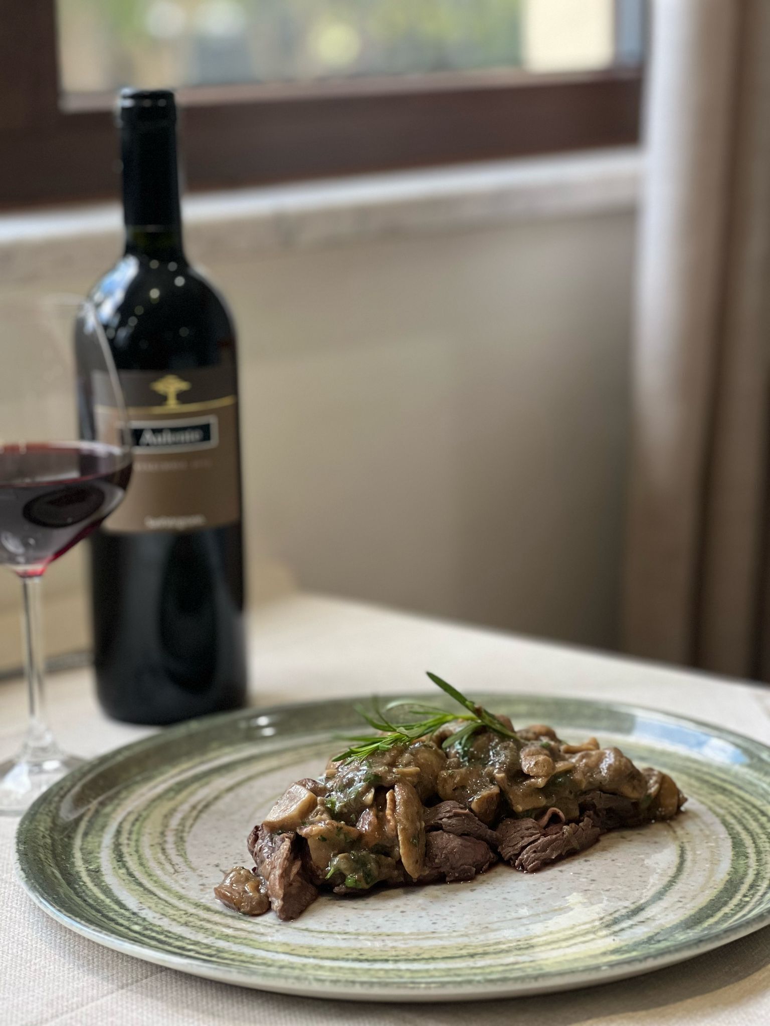 A plate of beef with mushrooms and a sprig of herb, a glass of red wine, and a bottle of wine on a table by a window.