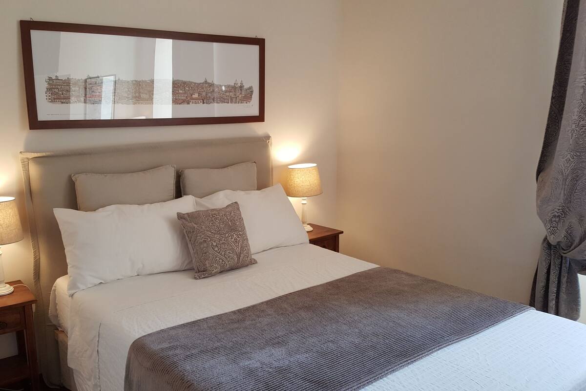 A neatly made bed with a patterned throw, two bedside lamps, and a framed cityscape picture above the bed.