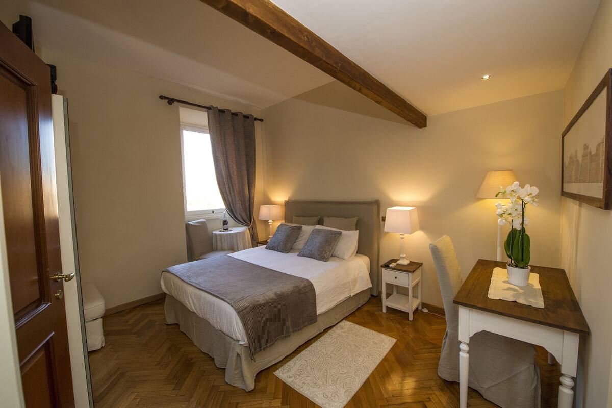 A bedroom with a large bed, bedside tables, a desk, a chair, a window with curtains, and a framed picture on the wall.
