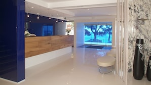 A modern reception area with a wooden counter, a seating area, and a view of trees through large windows. - Canyelles Platja (Roses)
